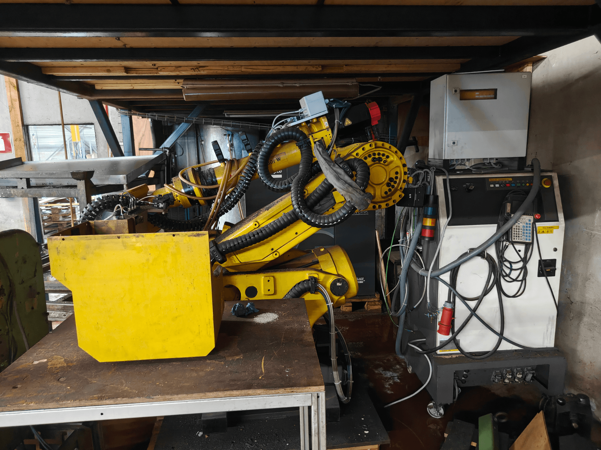Framifrånvy av en FANUC-robot S-430i W-arm, gul, med kontrollpanel, kablar och en gul komponent på en arbetsbänk.