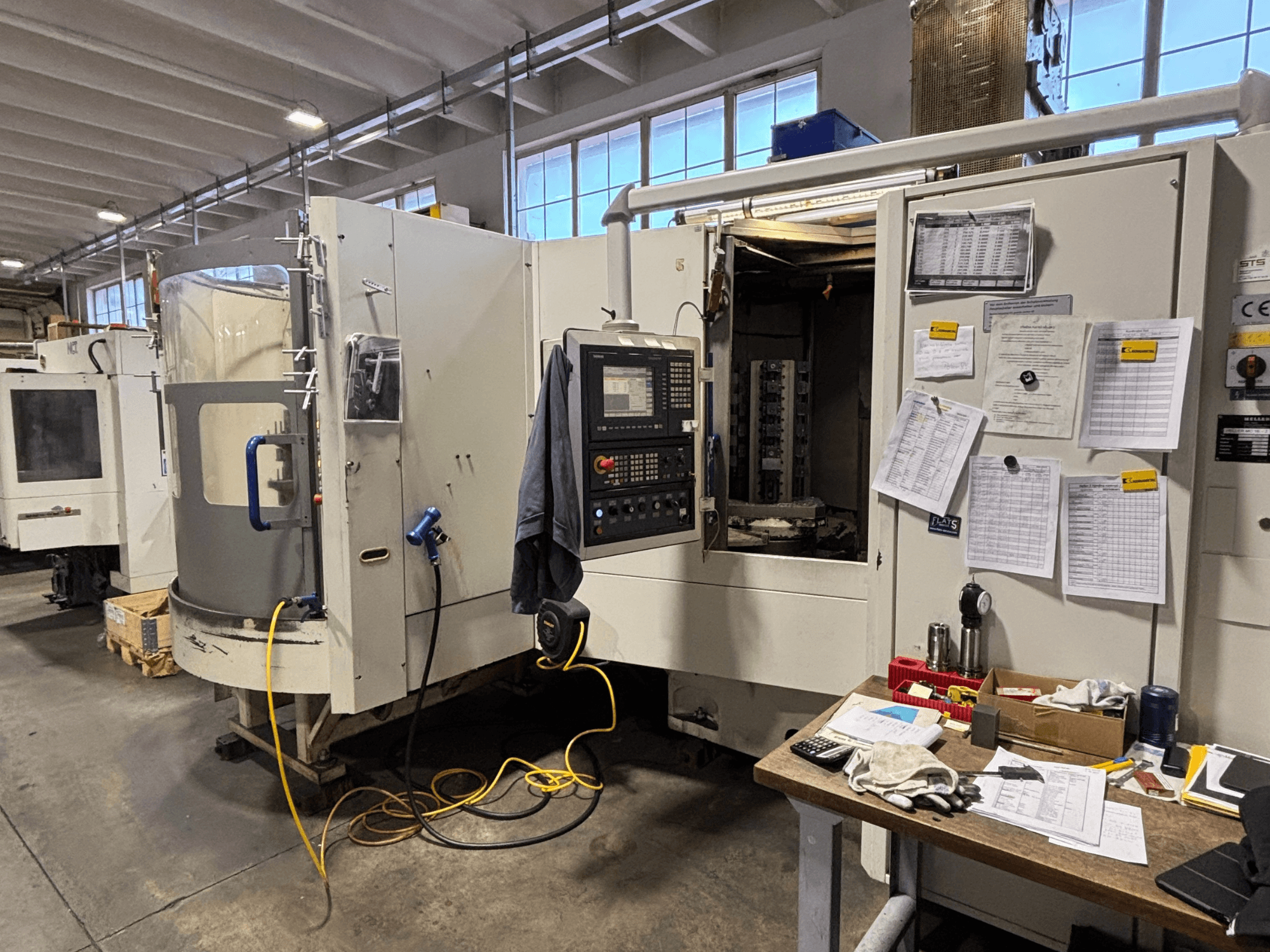 CNC-maskin i en verkstad, vy framifrån, med kontrollpanel och dokumentation på sidan; verktyg och material på bordet.