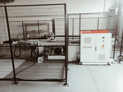 Vit CRIPPA-maskin bakom en säkerhetsbur, med synliga slangar och komponenter, vy från sidan, industrimiljö.