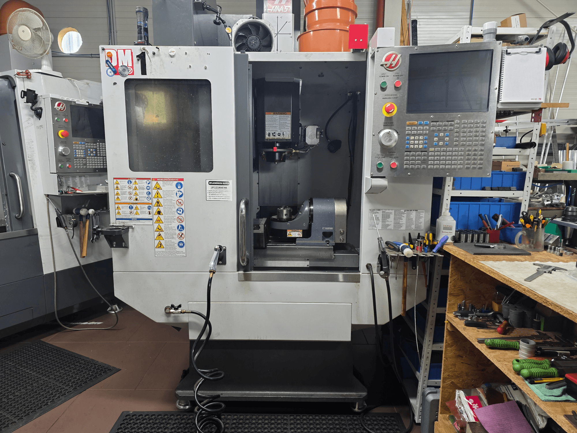 Haas CNC-maskin modell VF-2, vy framifrån, med reglage, spindelområde, säkerhetsetiketter och verktyg på en närliggande arbetsbänk.