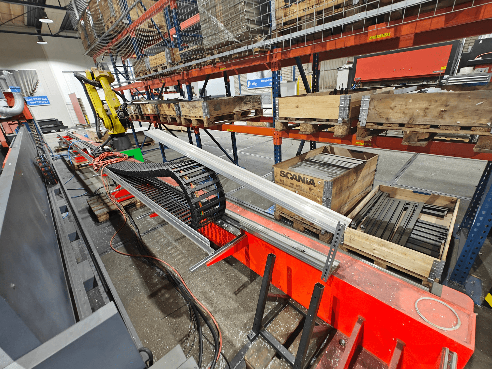 Robotarm på arbetsstation, orange och svarta kablar synliga, med Scania-lådor och aluminiumprofiler i en fabriksmiljö.