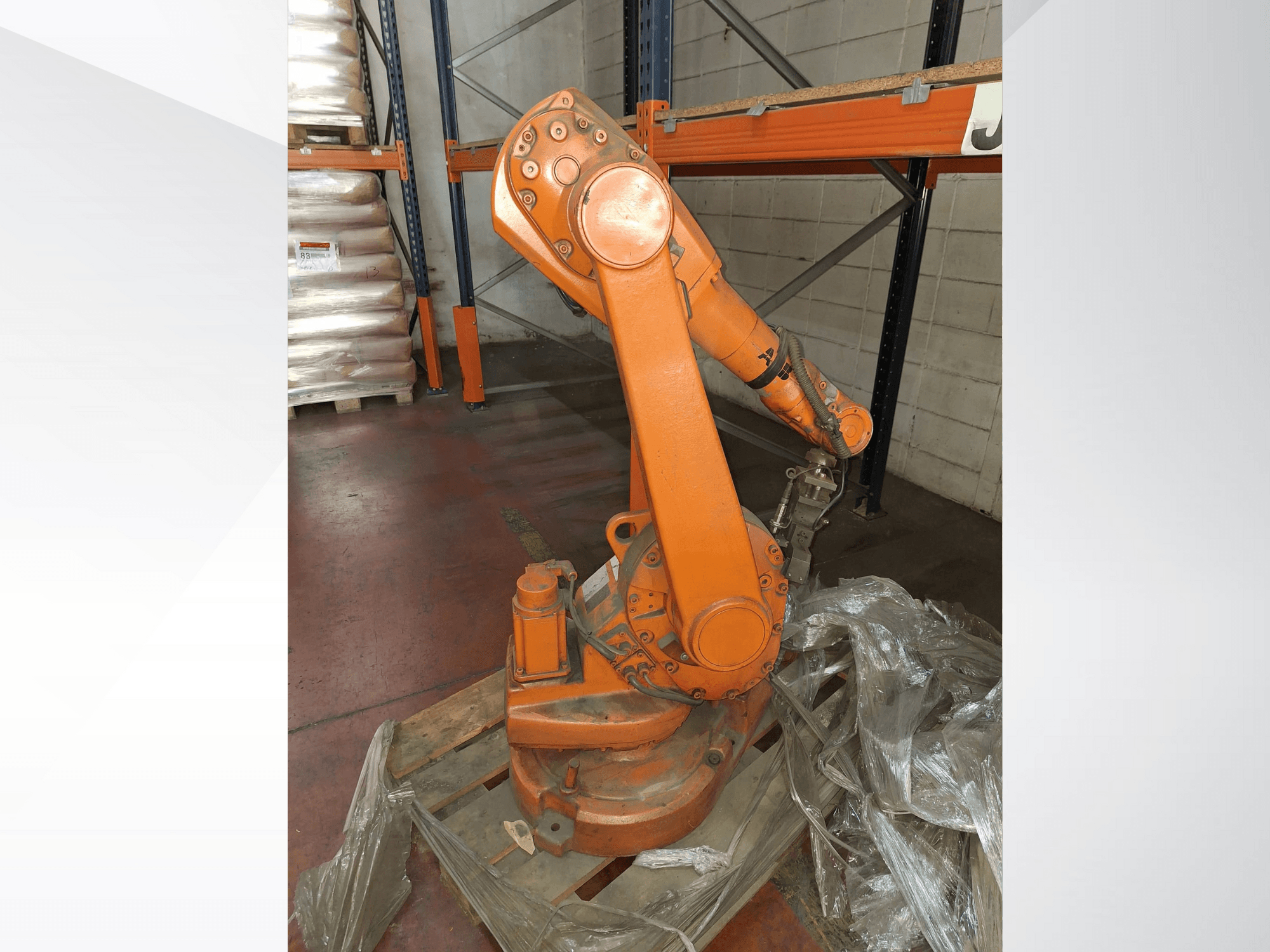 Industriell robotarm i orange, framifrån på träpall, med ansluten kabel och skyddande plastfolie runt basen.