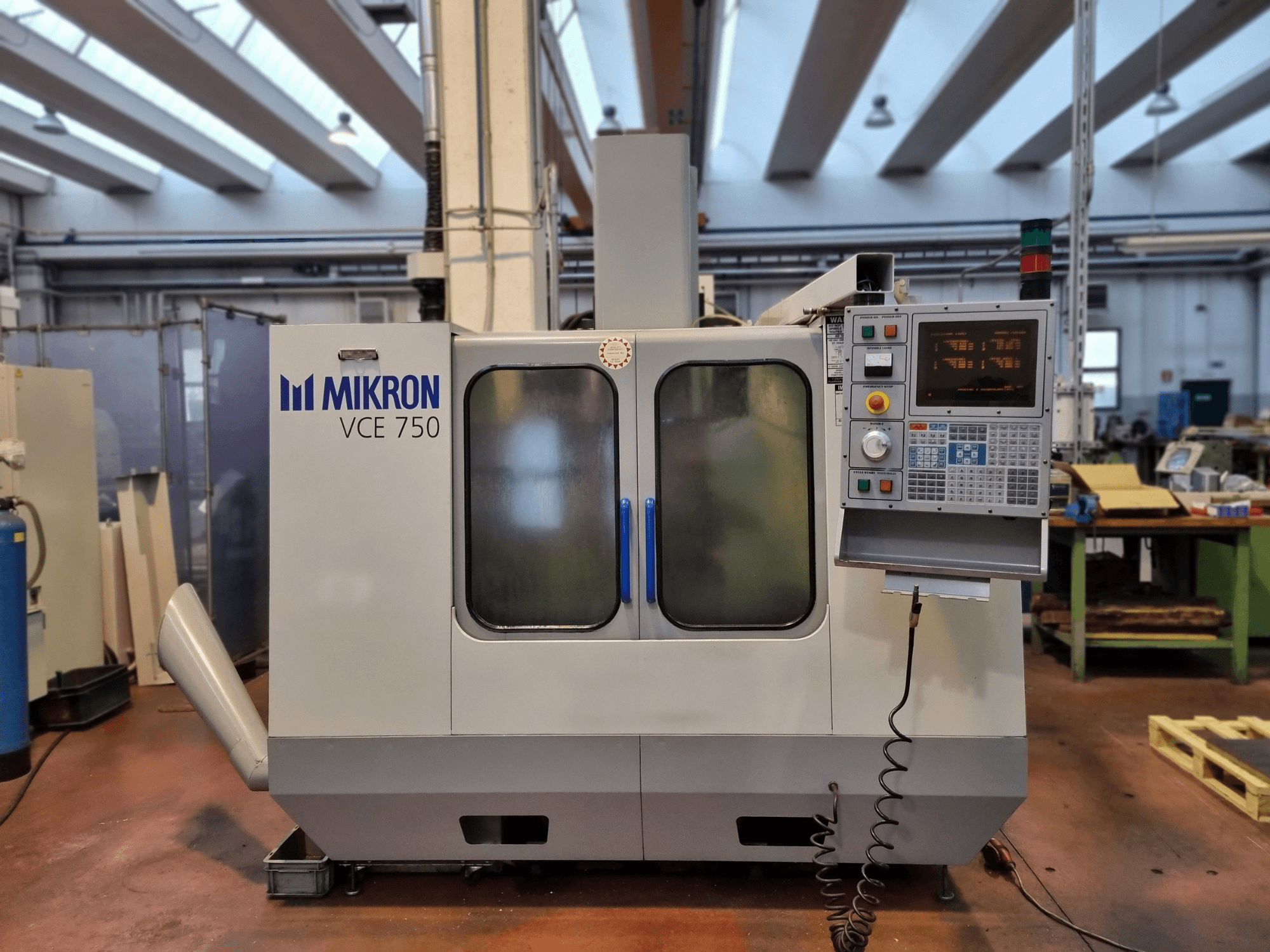 Vit Mikron VCE 750 CNC-maskin i en verkstad, vy från sidan, med digital kontrollpanel och stora glasdörrar.
