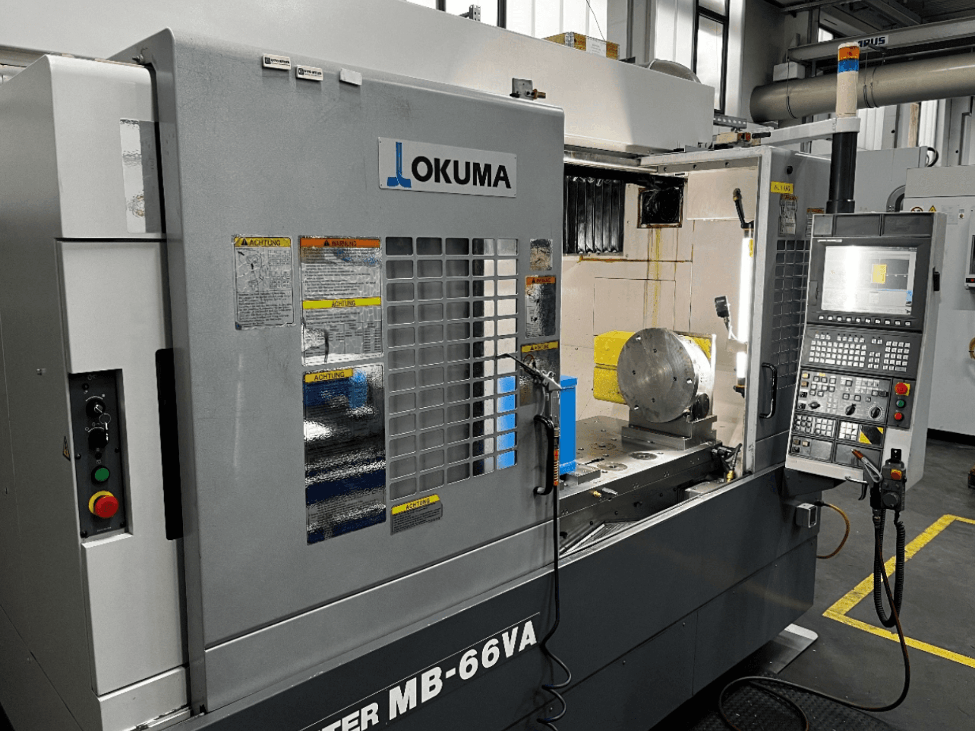 Okuma MB66-VA vertikal fleroperationsmaskin framifrån med kontrollpanel, arbetsstycke och maskininredning med säkerhetsetiketter.