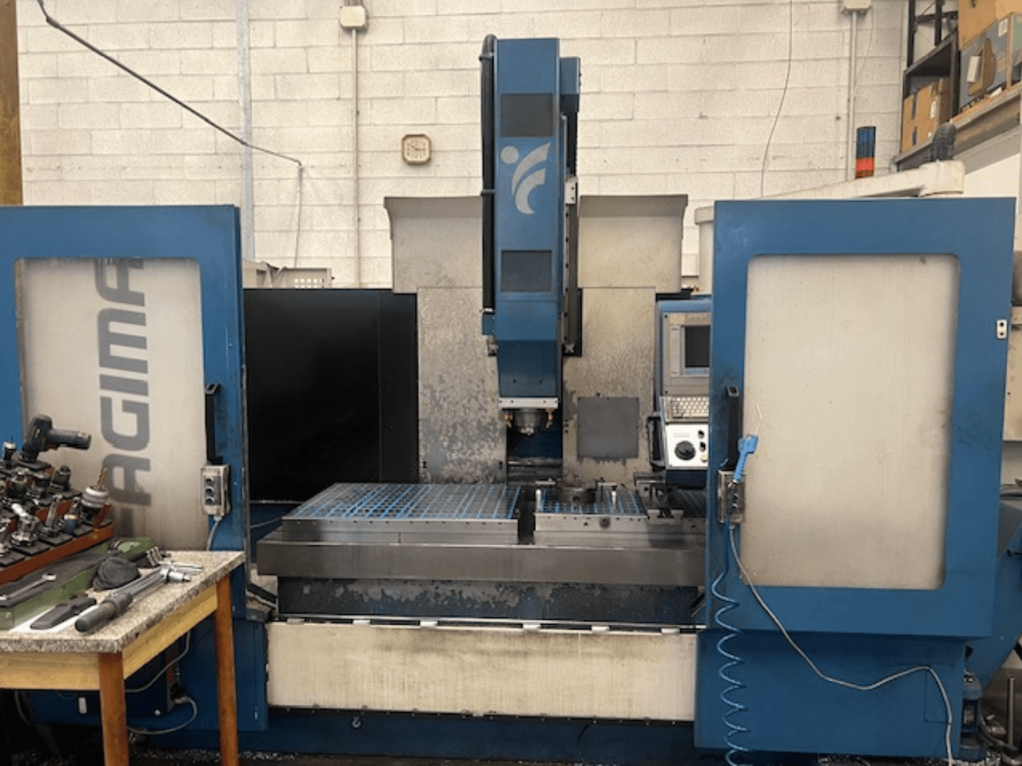 En frontvy av en JAGIMA CNC-maskin med en stor arbetsyta i metall, synlig kontrollpanel och verktyg på ett bord i närheten.