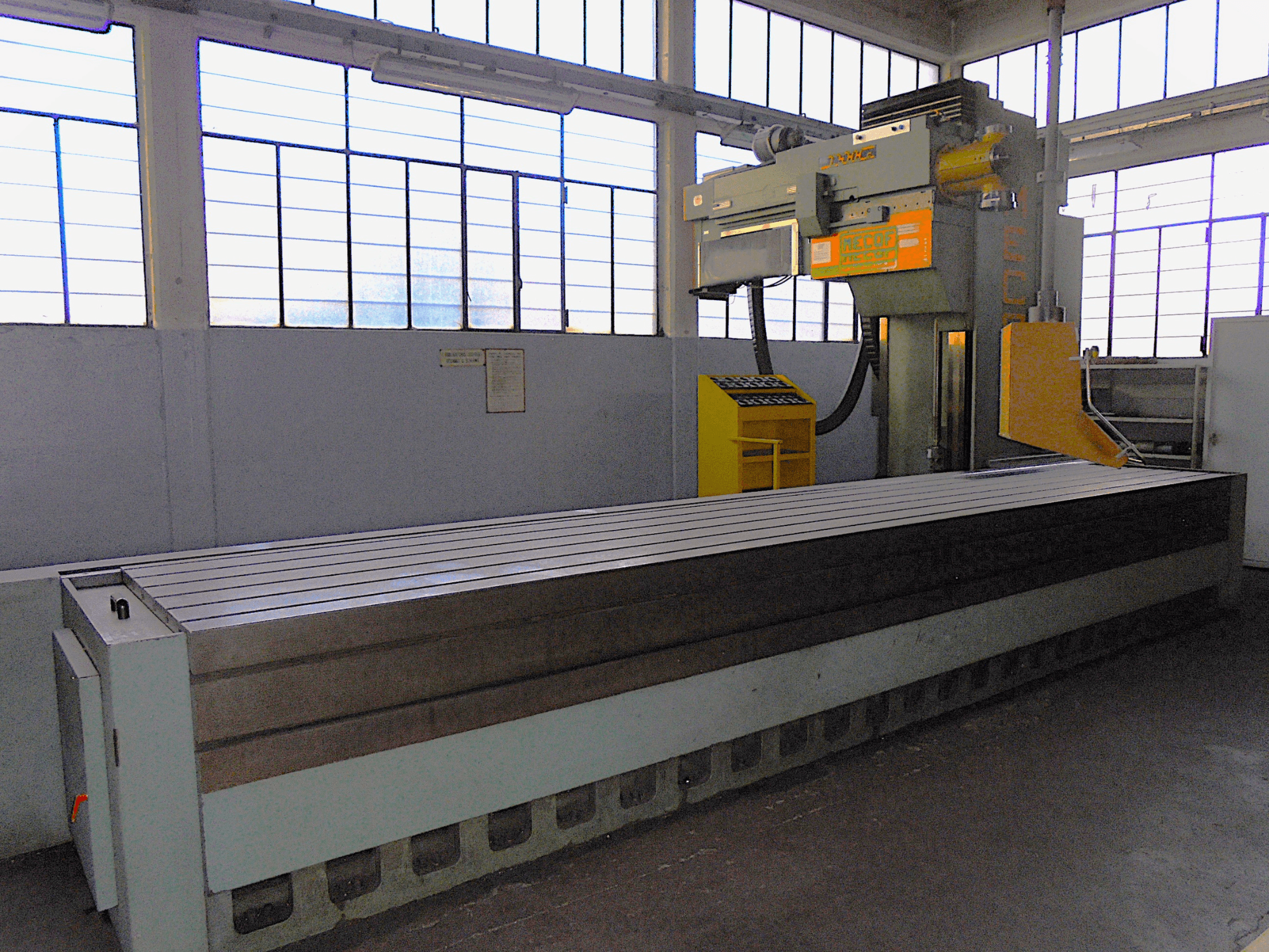 Vertikal fräsmaskin av okänt märke i en fabrik, vy från sidan, med ett stort bord med räfflad yta och kontrollpanel.