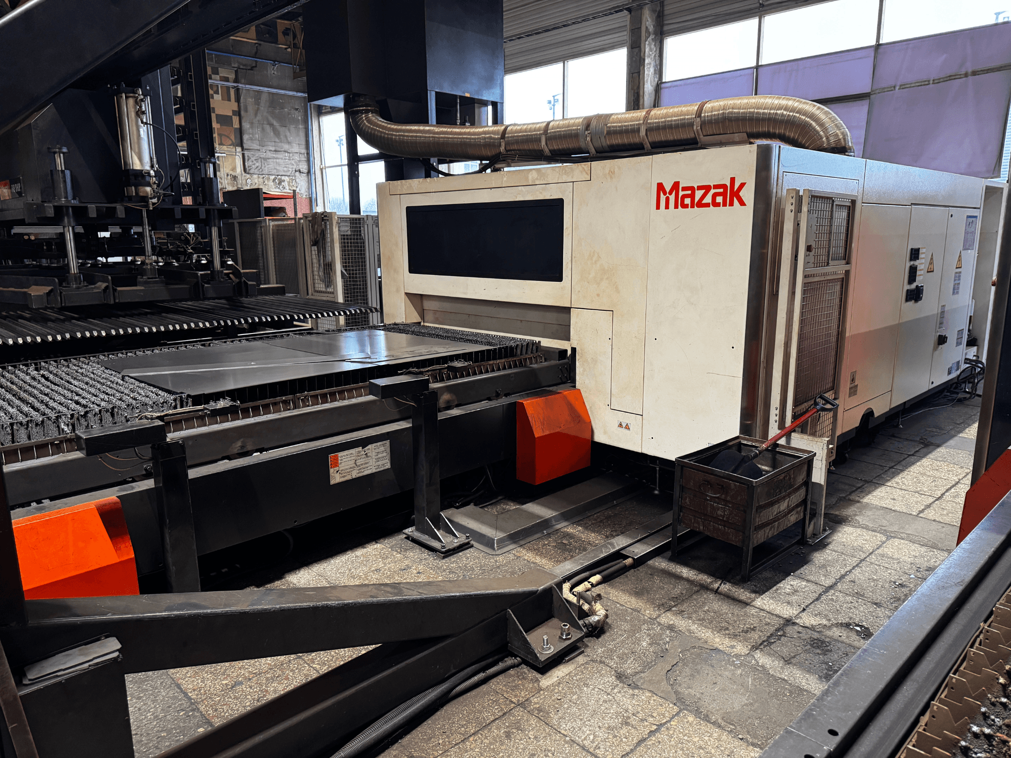 Mazak laserskärmaskin i en verkstad, sidovy som visar kontrollpanel, transportör och en uppsamlingstunna.
