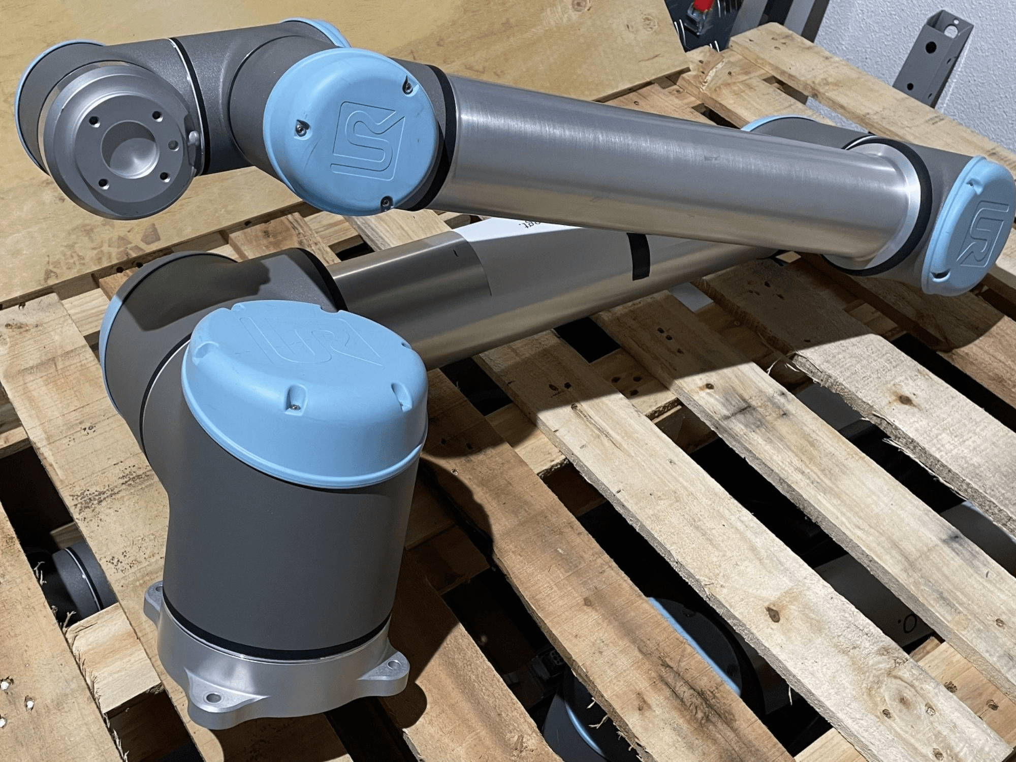 Silverfärgad robotarm med blå ändstycken, framifrån, med leder och mekaniska detaljer, vilande på en träpall.