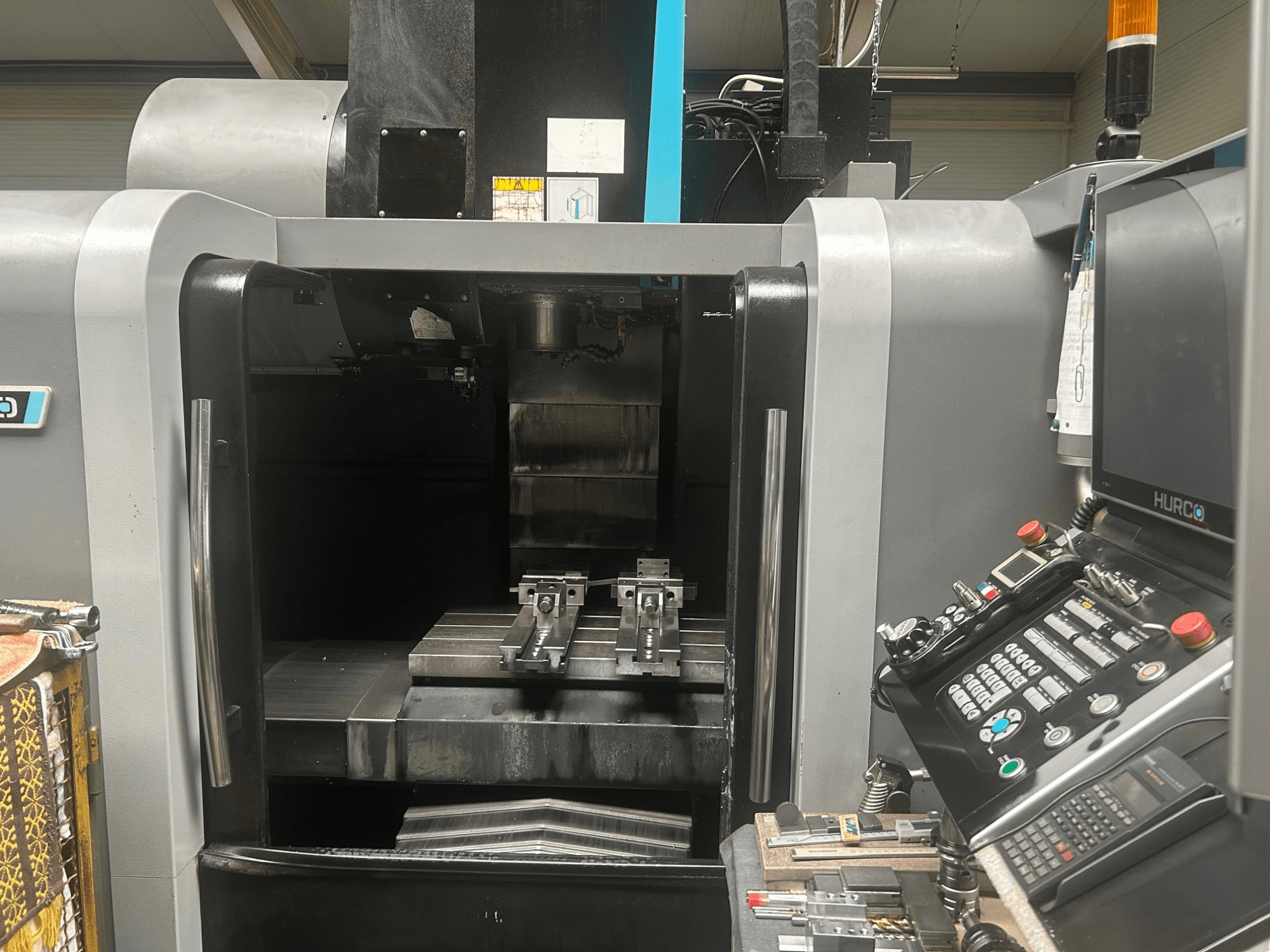 Hurco CNC-bearbetningscenter framifrån, med verktygshållare, arbetsstycke av metall och kontrollpanel med knappar och display.