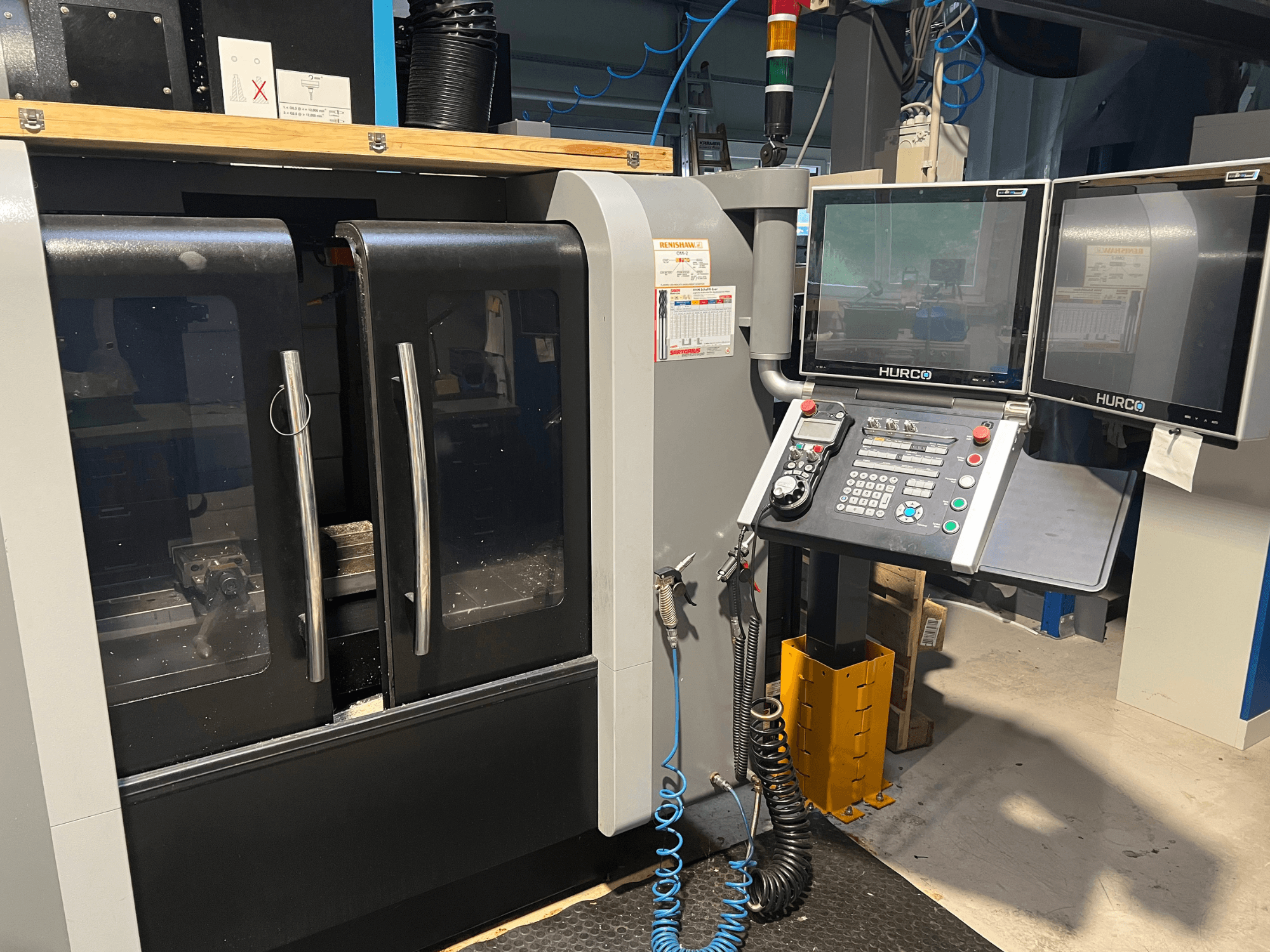 Hurco CNC-maskin framifrån med dubbla bildskärmar och kontrollpanel. Med genomskinliga dörrar och synligt bearbetningsområde på insidan.