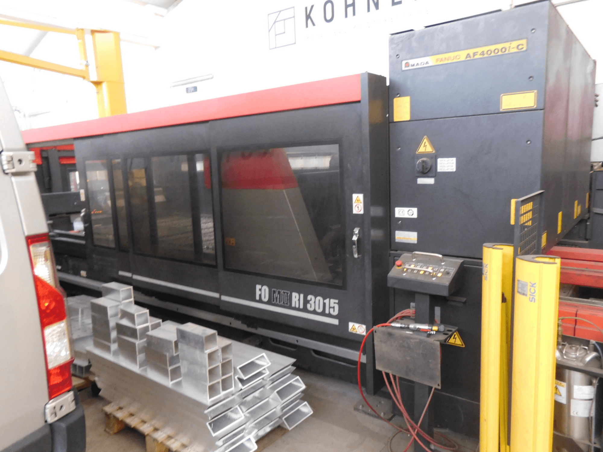 AMADA FO-3015 II RI CO2-laserskärmaskin i en verkstad, sidovy, med kontrollpanel och staplade aluminiumprofiler.