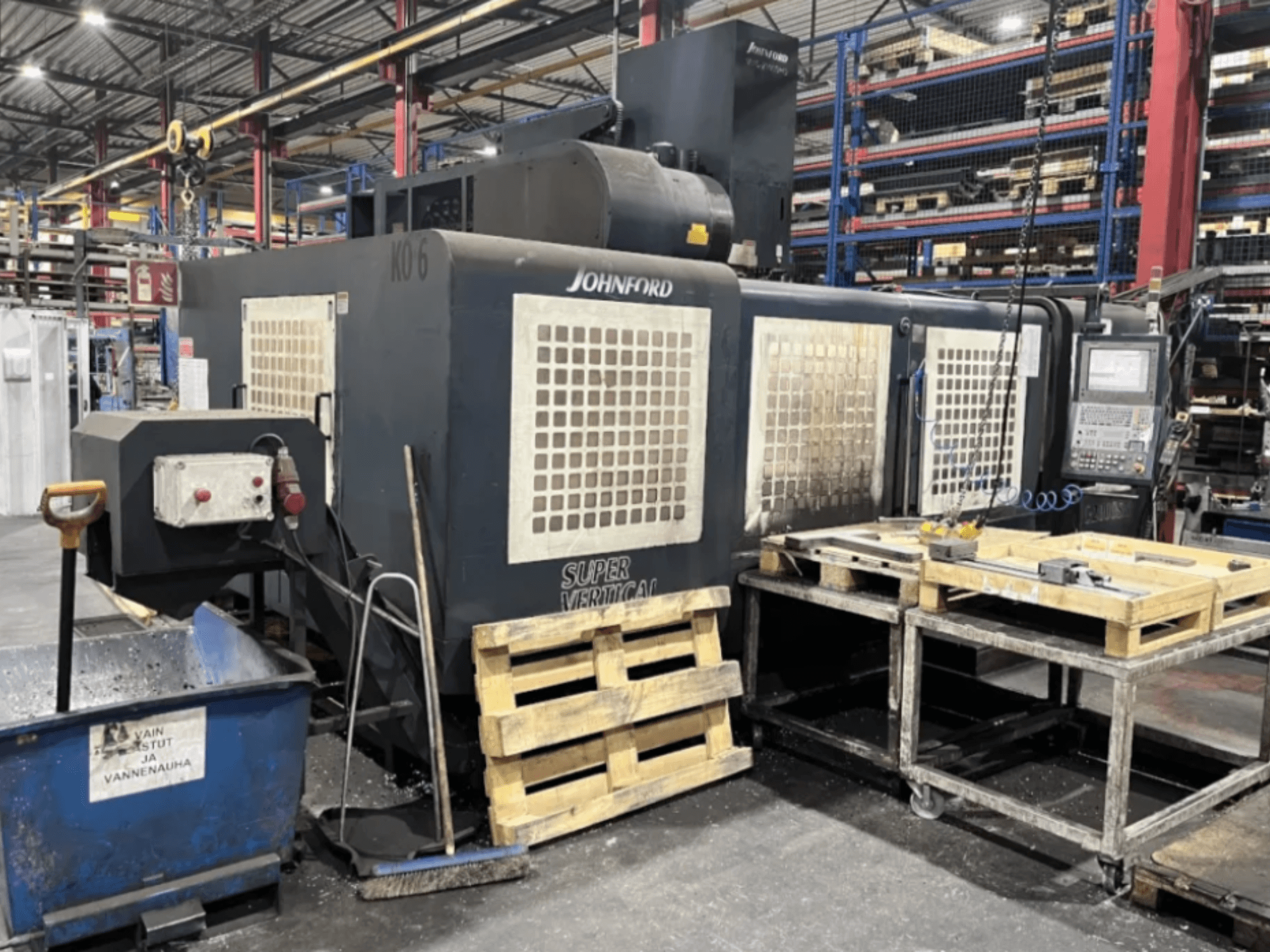 Johnford Super Vertical fräsmaskin i en fabrik, sidovy med kontrollpanel, pallar och rengöringsutrustning i närheten.