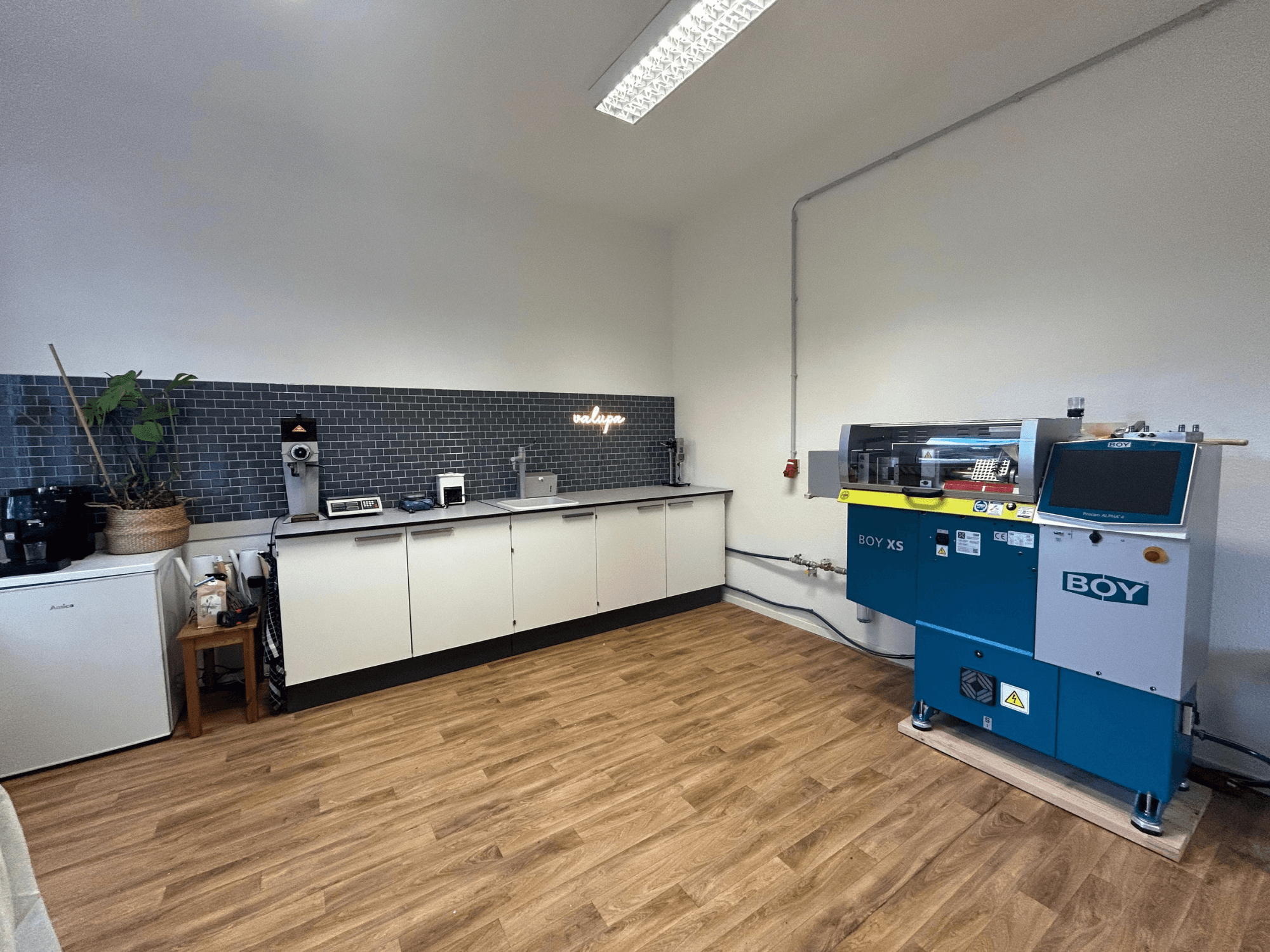 Hydraulisk formsprutningsmaskin BOY XS 161, framifrån, i en modern köksmiljö med vitvaror och trägolv.