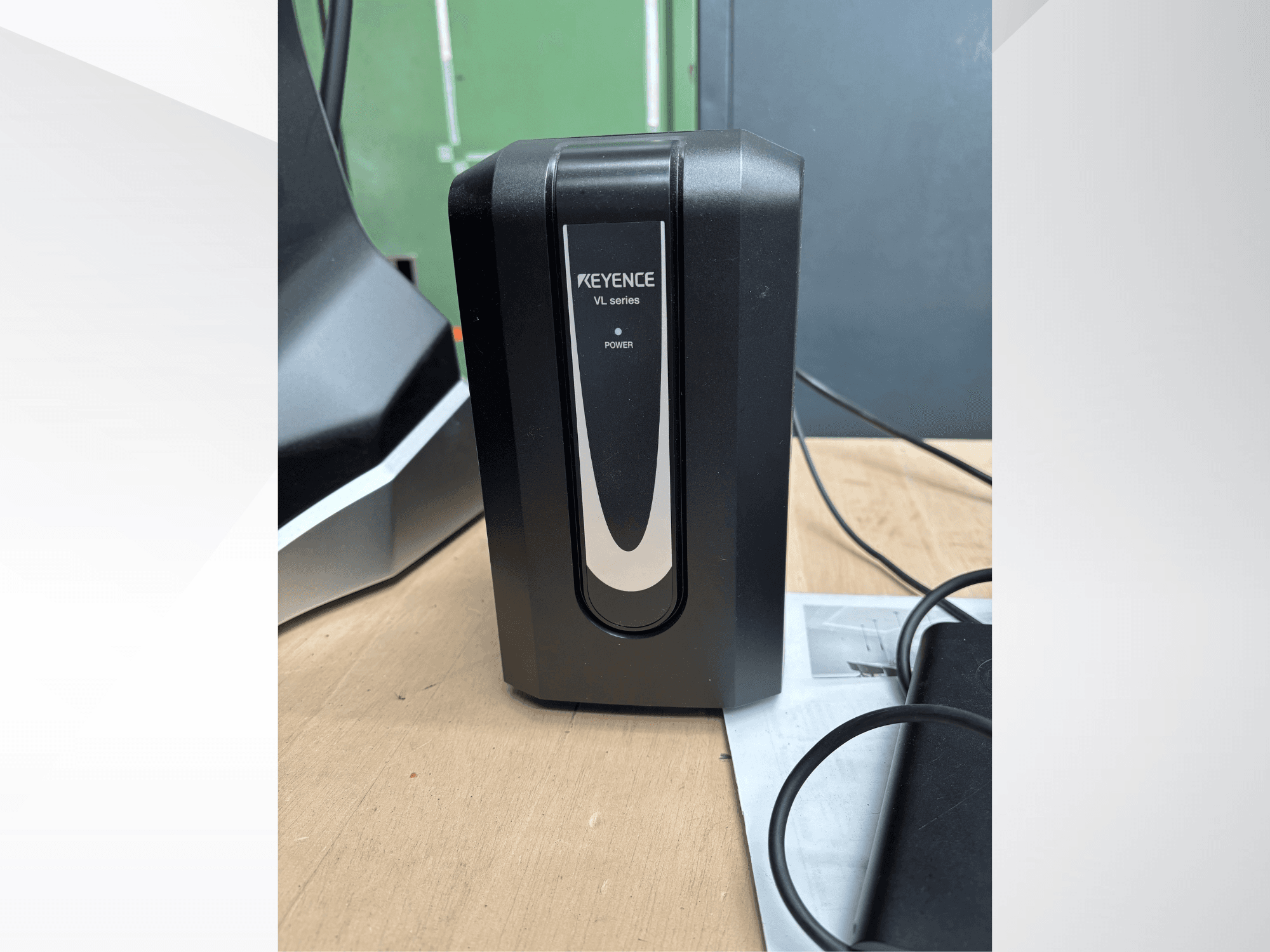 Svart strömförsörjningsenhet från KEYENCE VL-serien, sett framifrån, med strömindikator och anslutna kablar på en träyta.