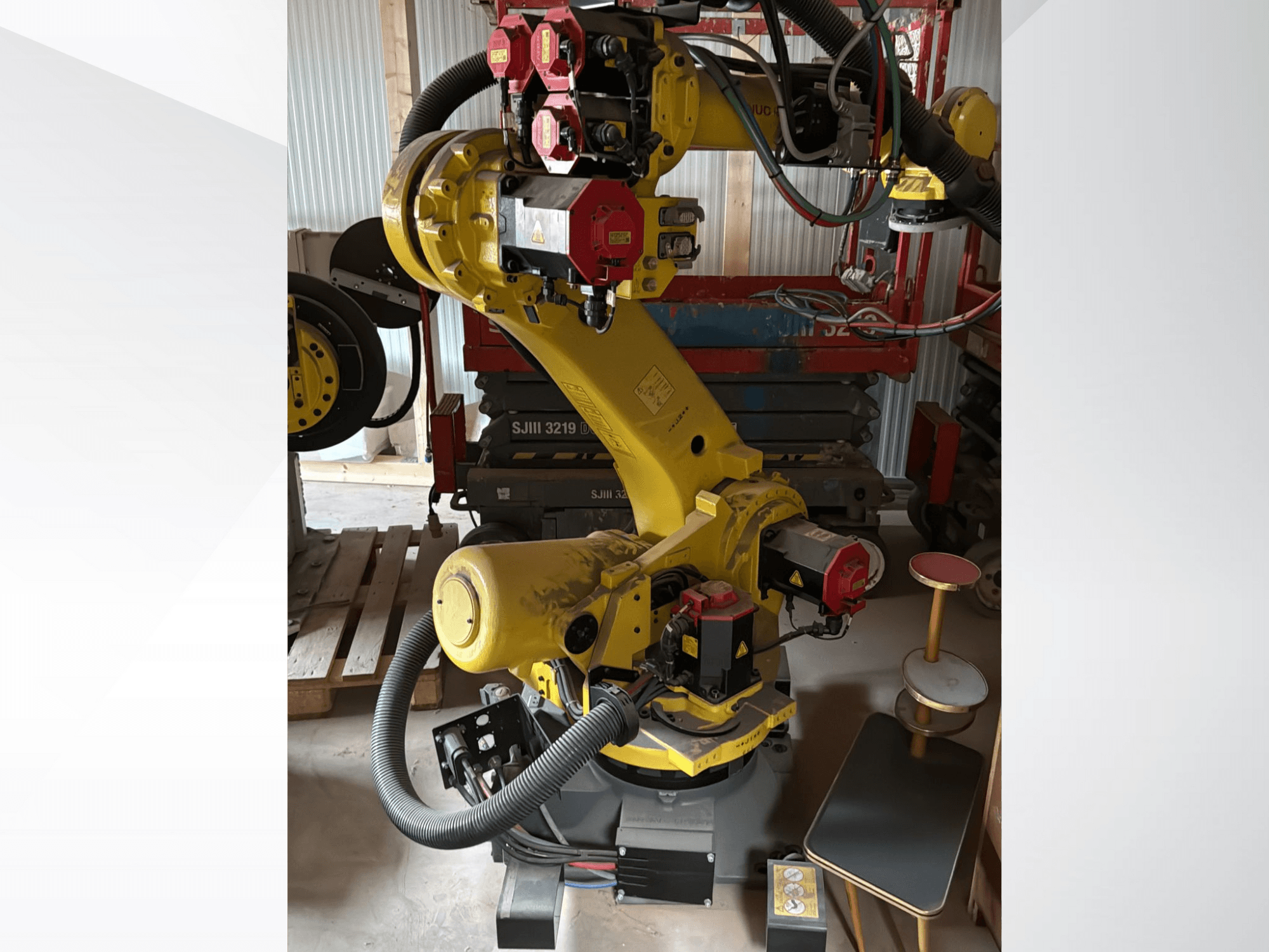 Gul industrirobotarm med röda styrenheter, framifrån; har flera leder och slangar, placerad på en arbetsplats.