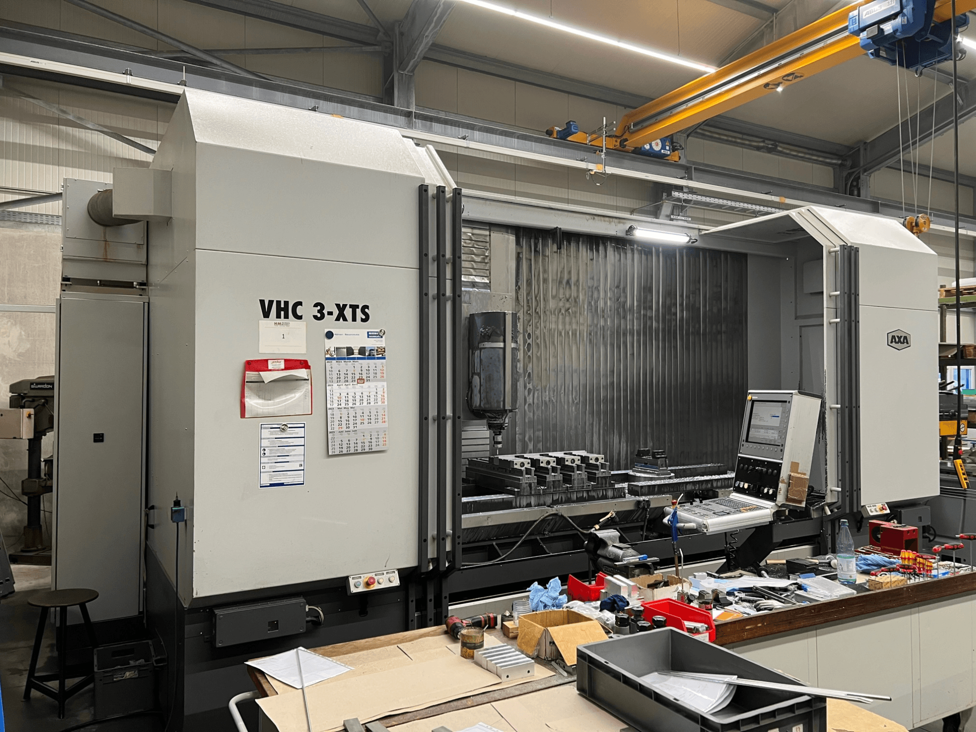 AXA VHC 3-XTS CNC-maskin i sidovy, med kontrollpanel och arbetsområde belamrat med verktyg och komponenter.