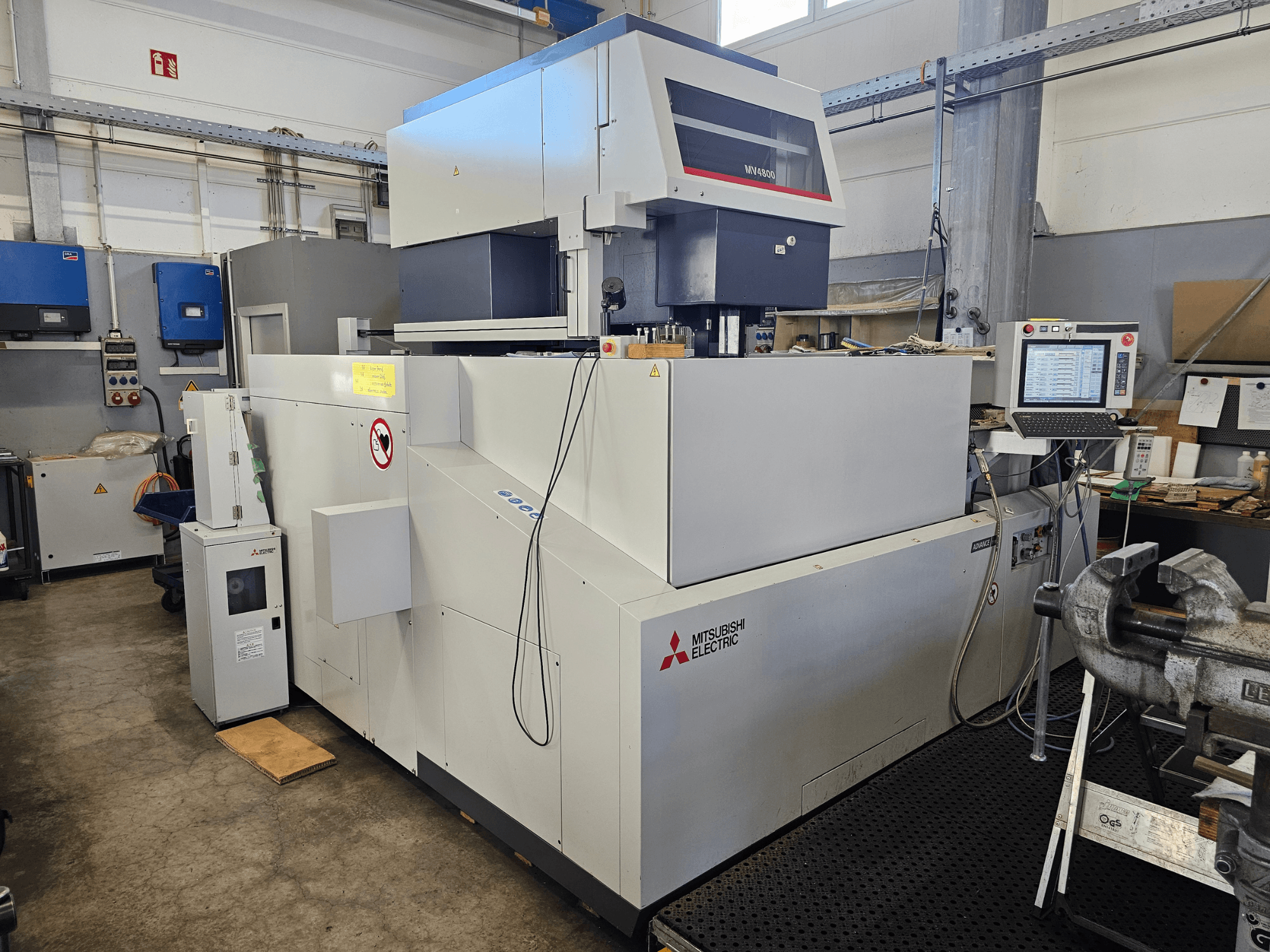 Mitsubishi MV4800 Wire EDM Machine i en verkstad, sidovy som visar kontrollpanel, säkerhetsetiketter och omgivande utrustning.