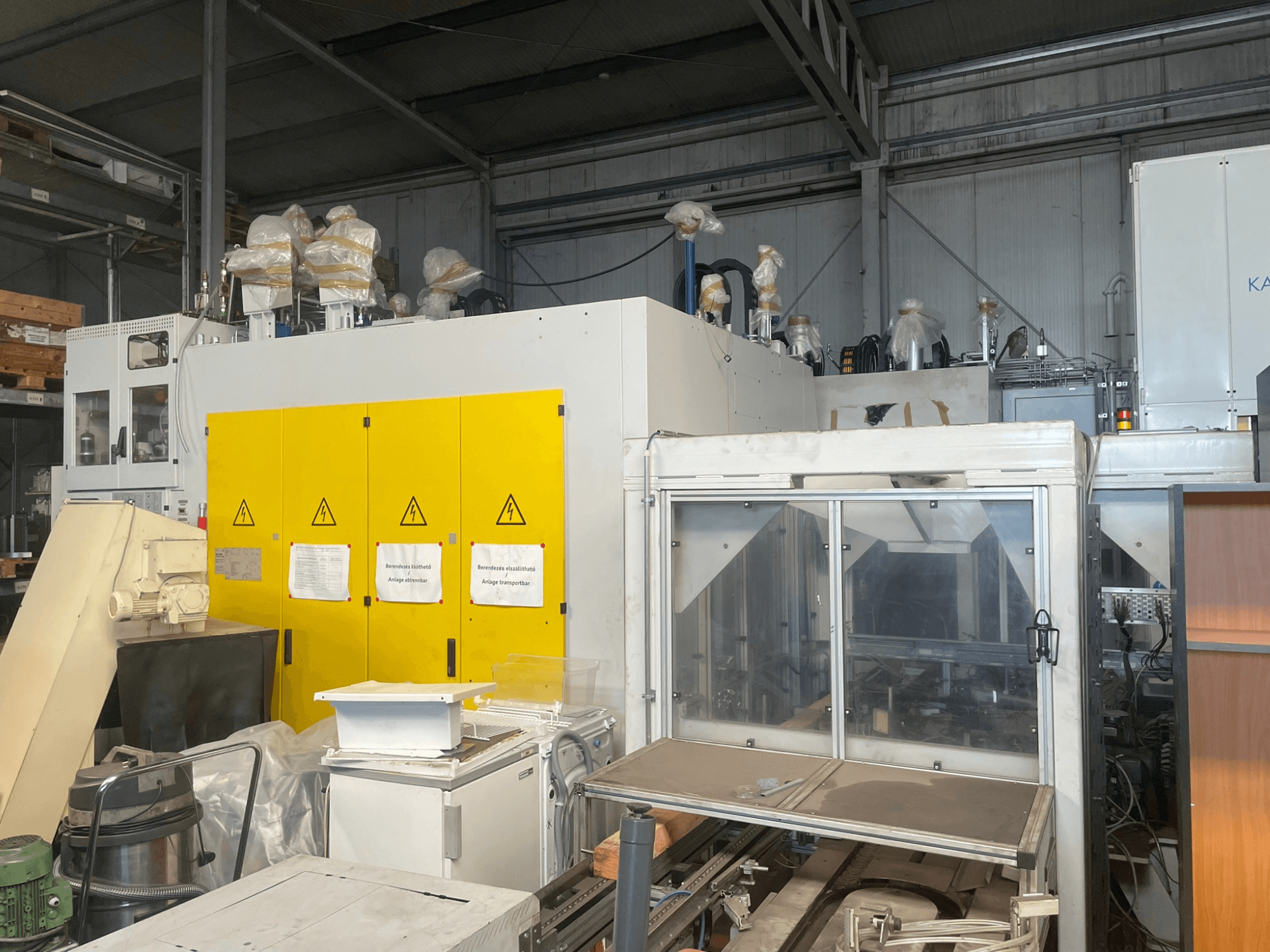 Industrimaskin med gul kontrollpanel, vy från sidan, med varningsetiketter och plastöverdrag, i en lagerlokal.