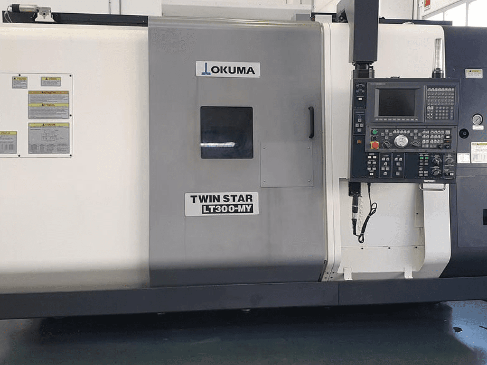 Okuma Twin Star LT300-MY CNC-svarv i sidovy, med kontrollpanel, manöverknappar och säkerhetsetiketter.
