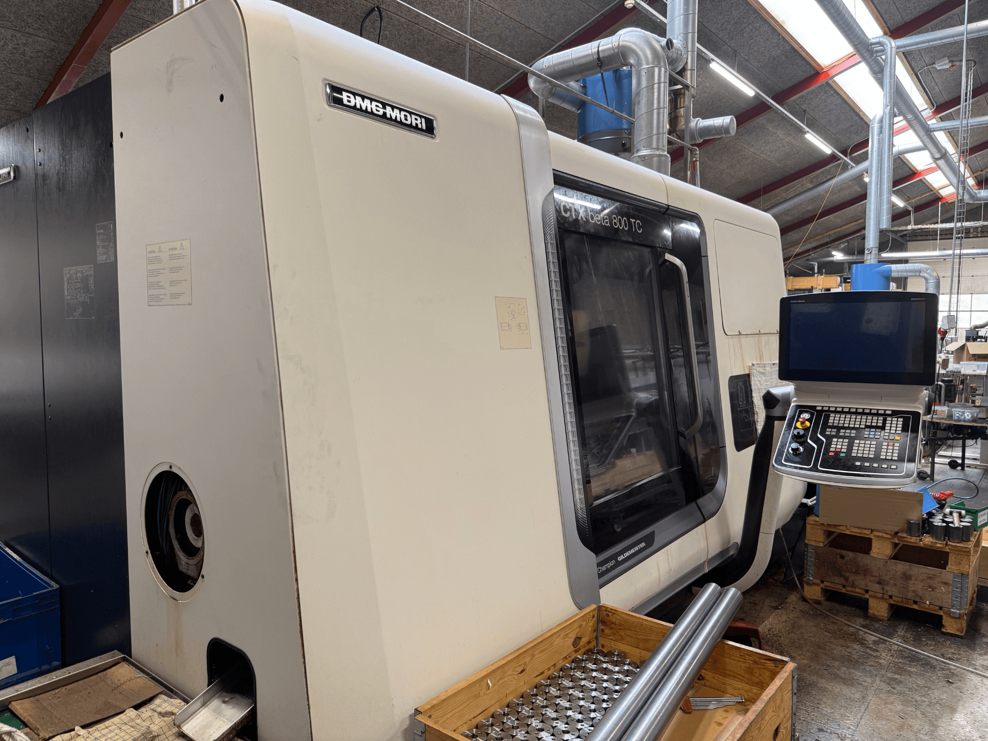 DMM-MORI CTX beta 800 TC-maskin, framifrån, med kontrollpanel, verktyg och metallstänger på en träpall.
