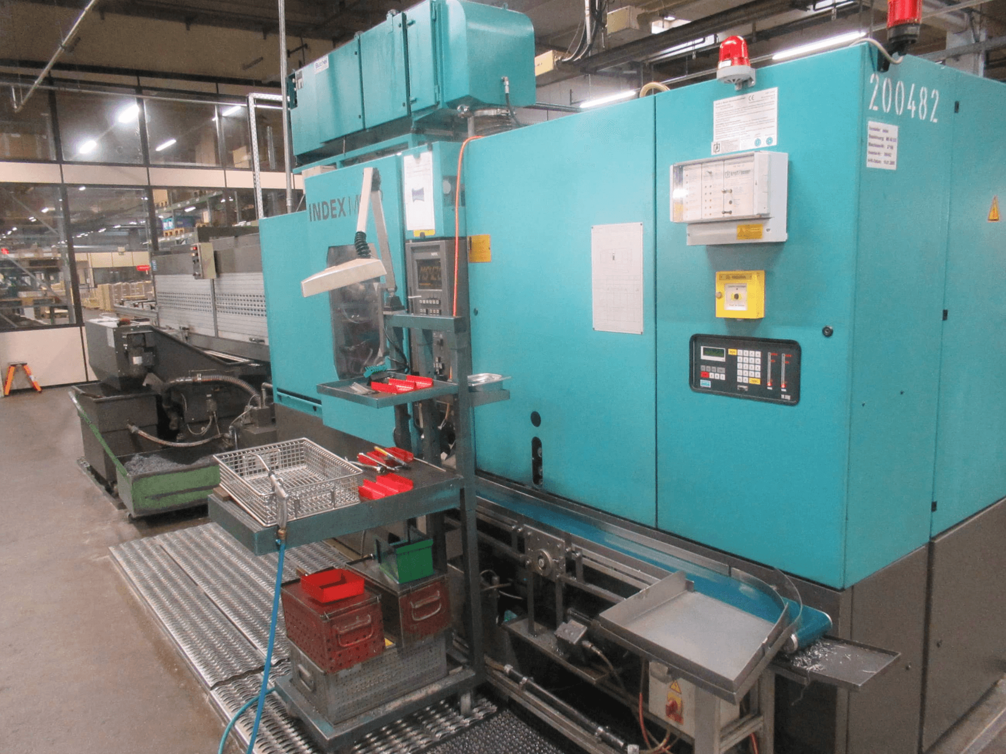 Turkos INDEX-maskin i fabrik, vy från sidan; har kontrollpanel, verktygsförvaring och transportör. Synliga fack, slangar och larm.
