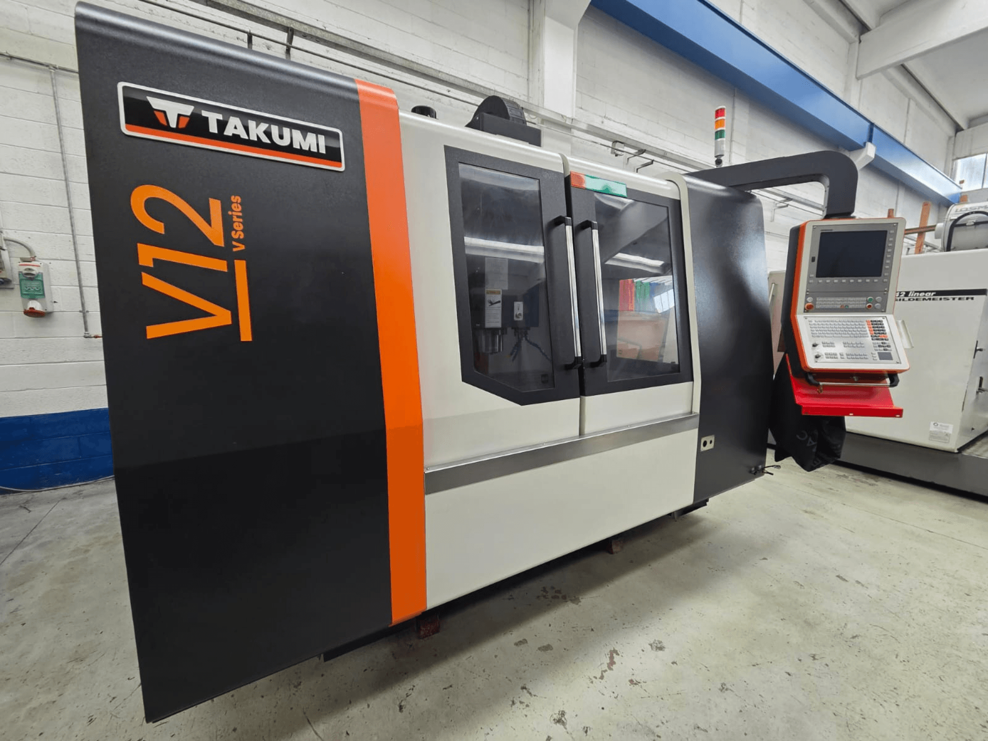 Takumi V12 CNC-fräsmaskin i V-serien, framifrån, har en kontrollpanel och stora glasfönster.