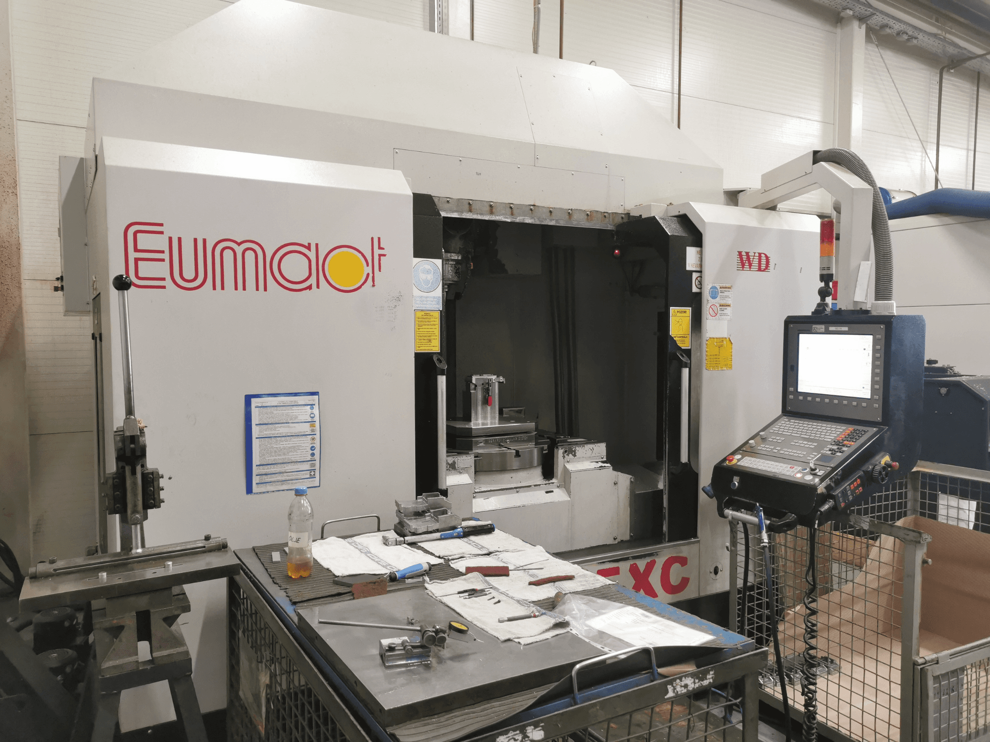 Frontvy av en Eumdaot CNC-maskin med kontrollpanel, verktyg och en arbetsyta full av komponenter och material.