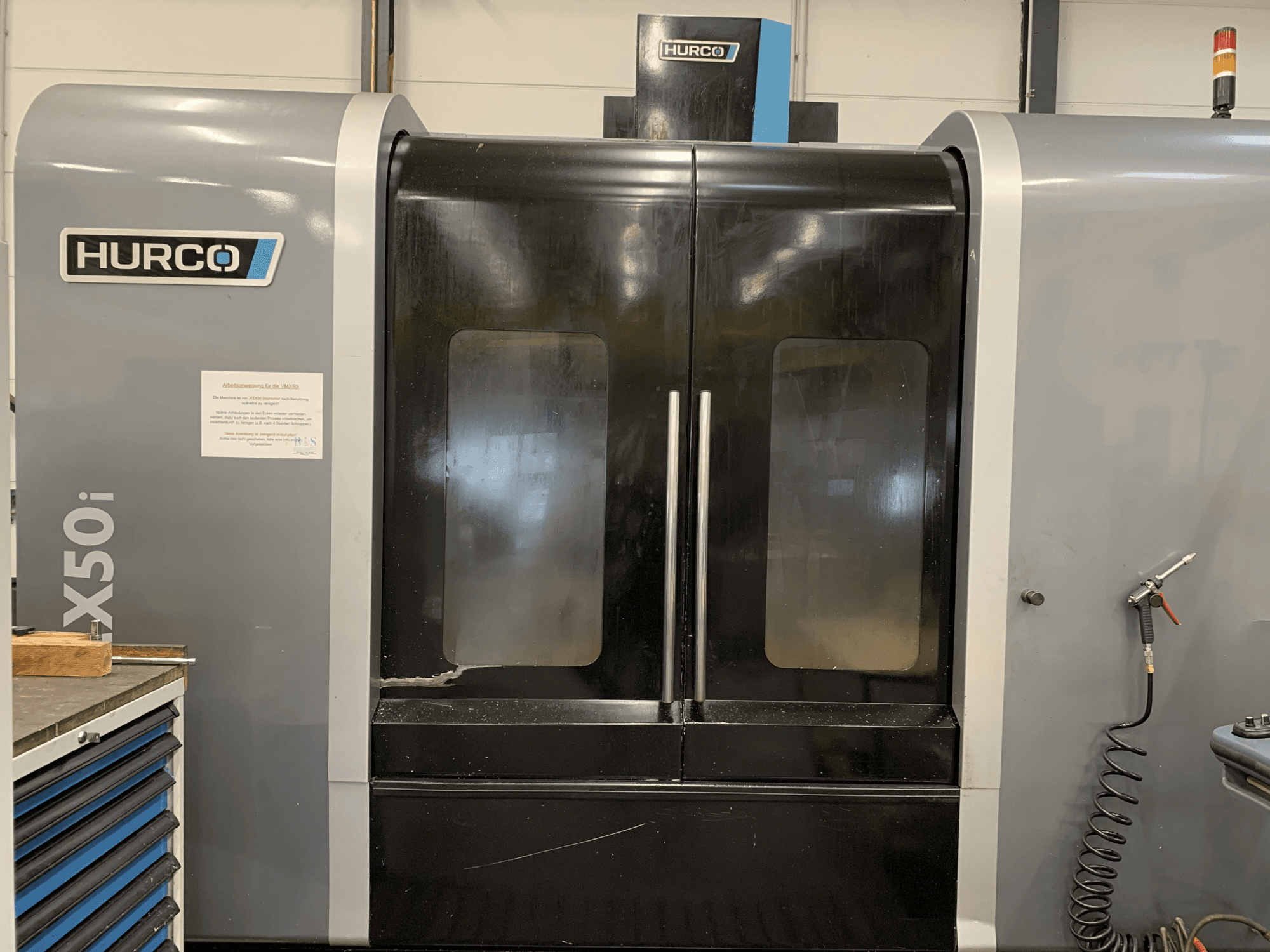 Frontvy av en Hurco VX50i CNC-maskin med dubbla dörrar, som visar tekniska kontroller och en underhållsskylt på sidan.