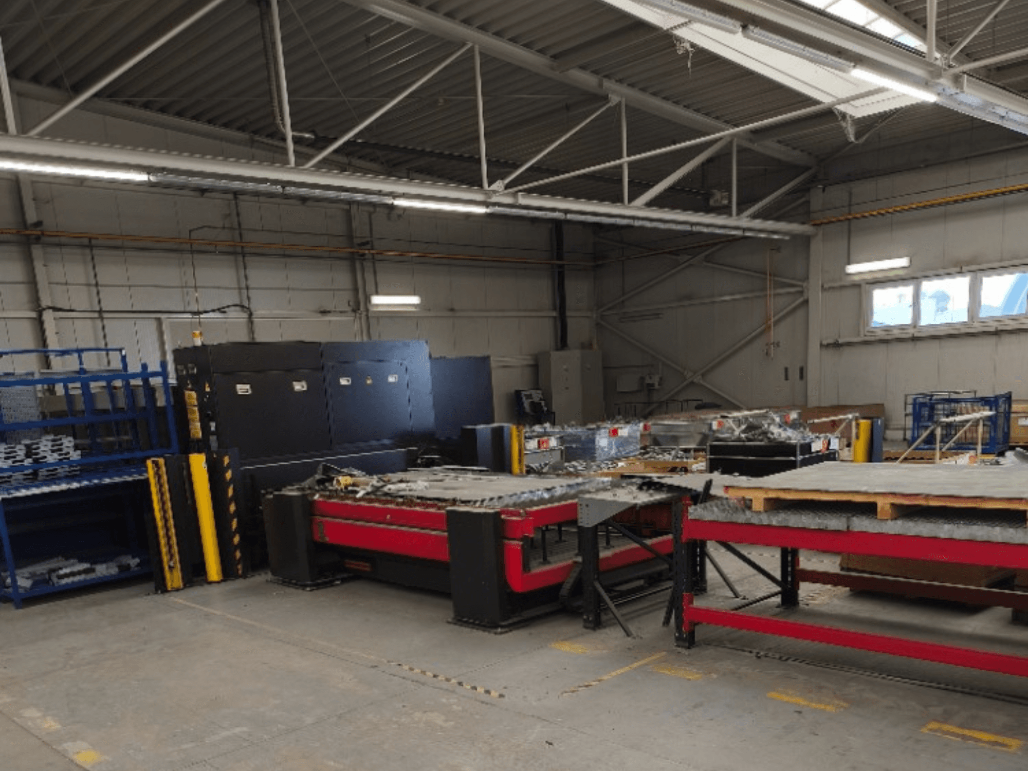 En AMADA LCG 3015 AJ fiberlaserskärmaskin i en verkstad, sedd framifrån med förvaringshyllor och material i bakgrunden.