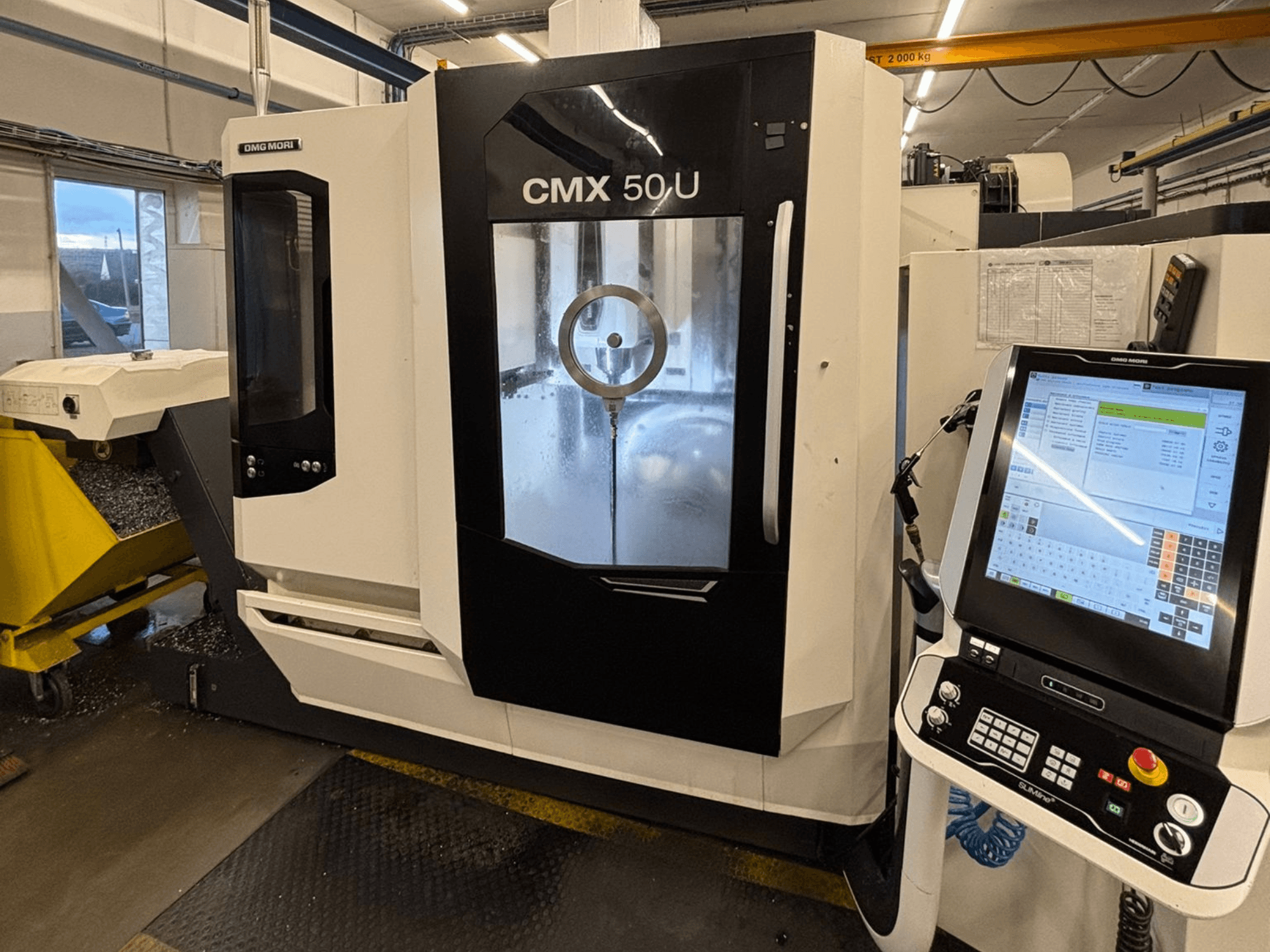 DMG MORI CMX 50 U vertikal fleroperationsmaskin, framifrån med kontrollpanel och glasskärm, som visar den inre mekaniken.