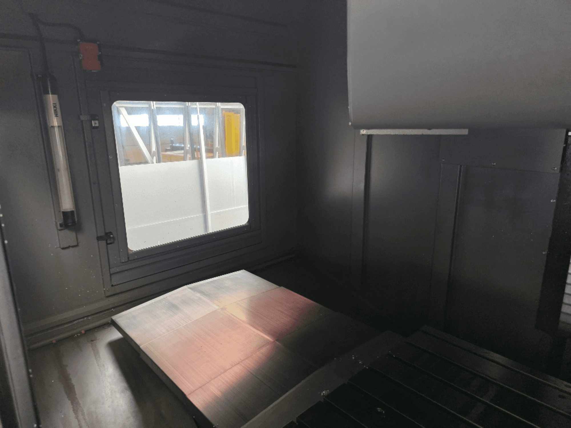 Invändig vy av ett maskinskåp med en polerad metallplatta, ett sidofönster och minimal belysning. Inget varumärke synligt.