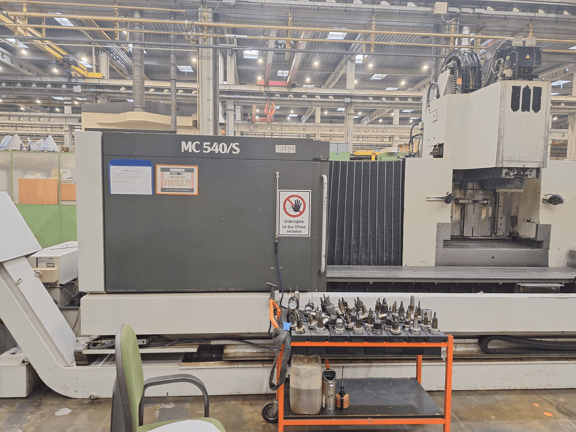 Vertikal bearbetningscenter STAMA MC 540S i en verkstad, sidovy med verktygshållare på en vagn i närheten.