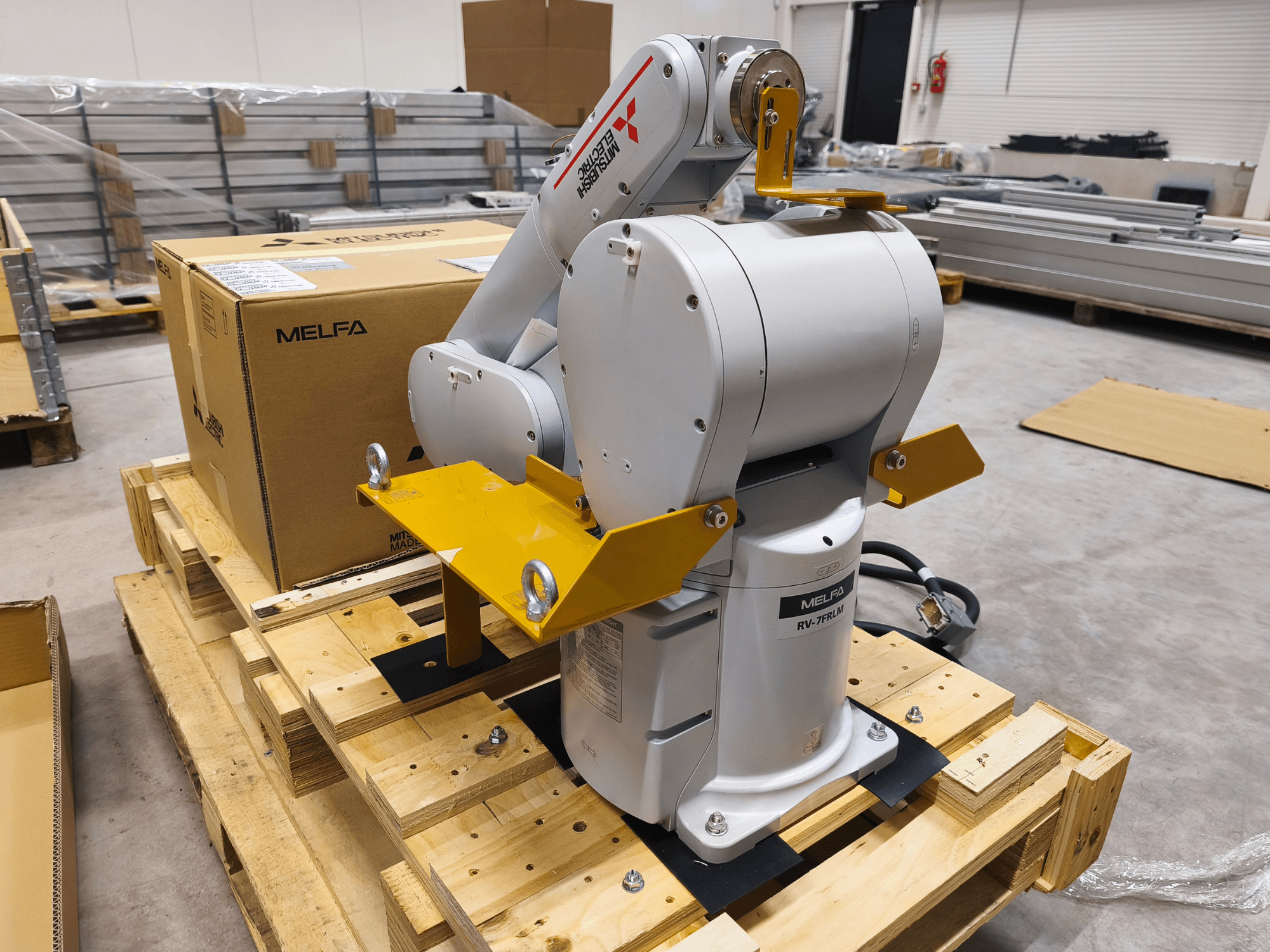 MELFA RV-7FLM industriell robotarm på träpall, framifrån, med gula stödfästen och grå förpackningslåda.