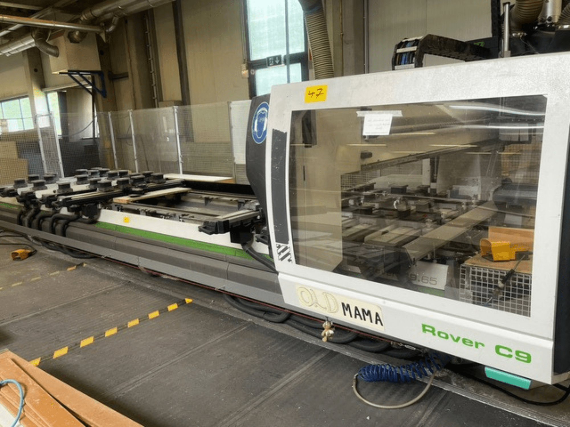 BIESSE Rover C 9.65 CNC-fräs i en verkstad, sidovy som visar kontrollpanelen och arbetsbordet med klämmor.