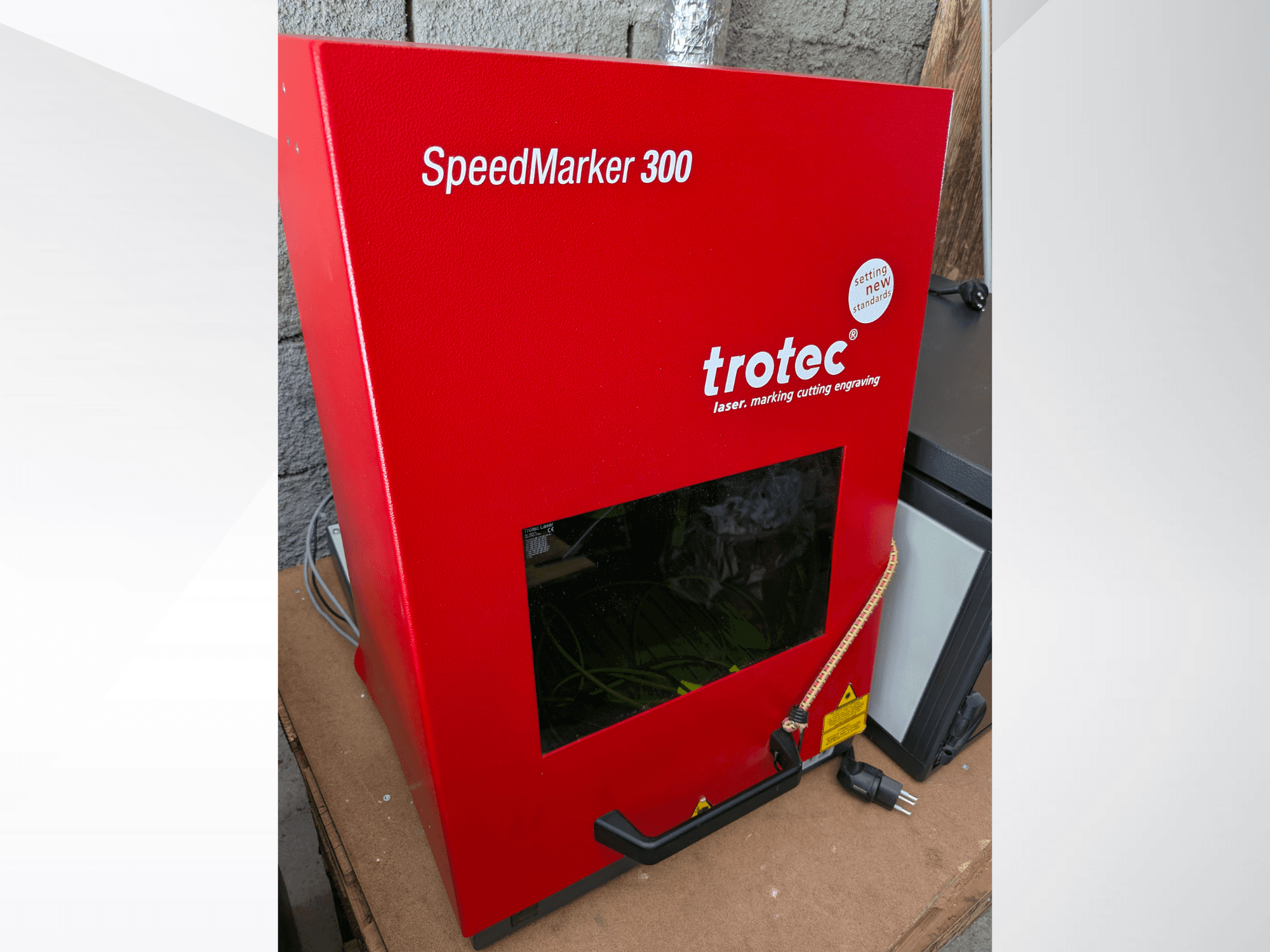 Röd Trotec SpeedMarker 300 lasermärkningsmaskin framifrån, med ett stort visningsfönster och strömanslutning.