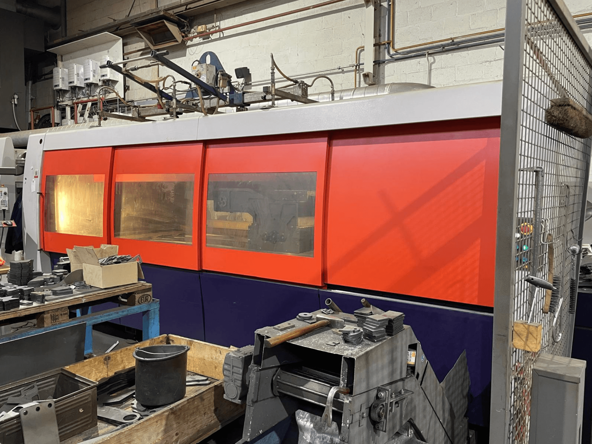 Närbild av en orangevit industriell laserskärmaskin, vy från sidan, med kontrollpanel och verkstadsverktyg i förgrunden.