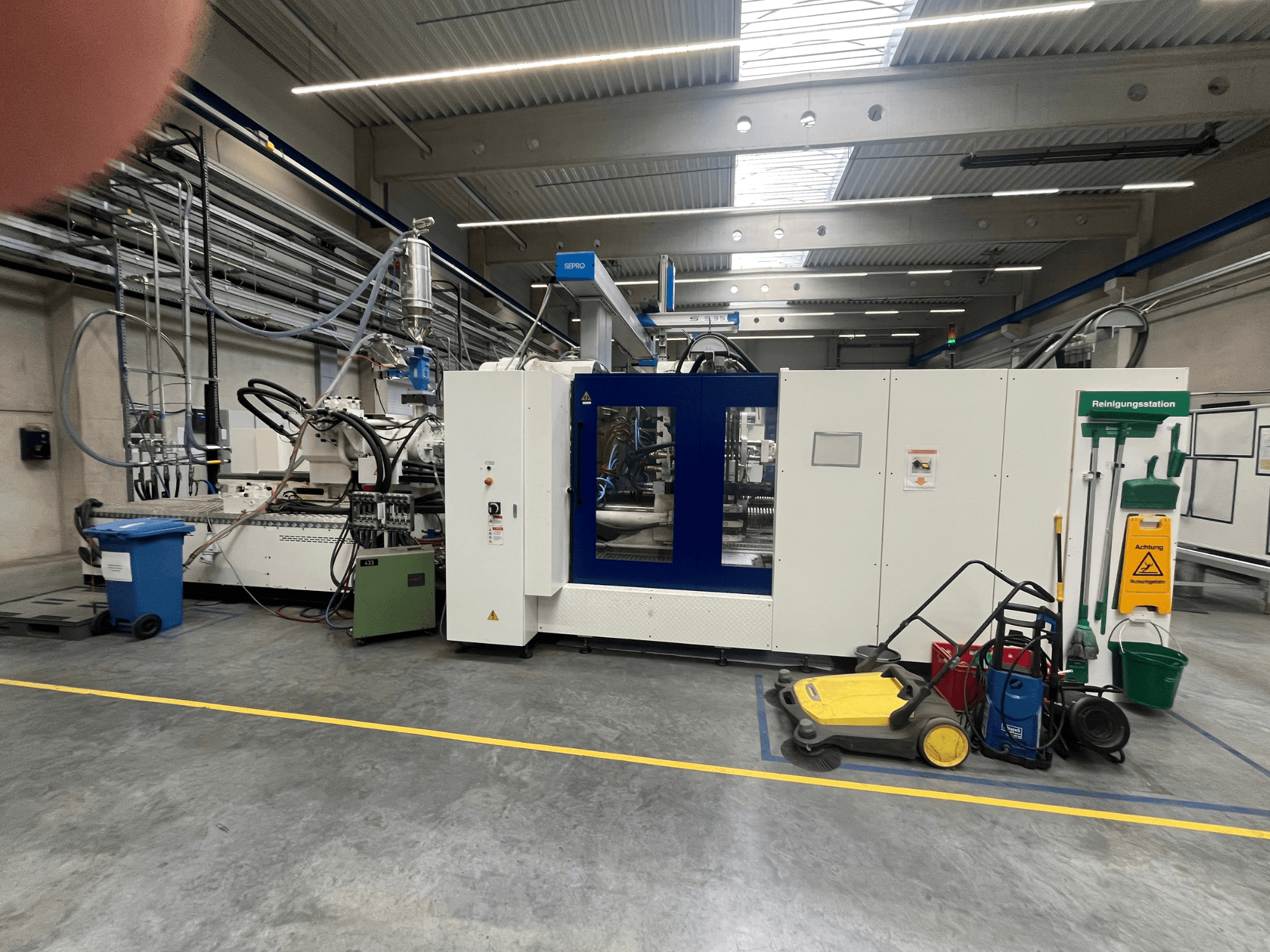 Industrimaskin i en fabrik, framifrån, med blå och vita paneler, slangar och rengöringsutrustning i närheten.