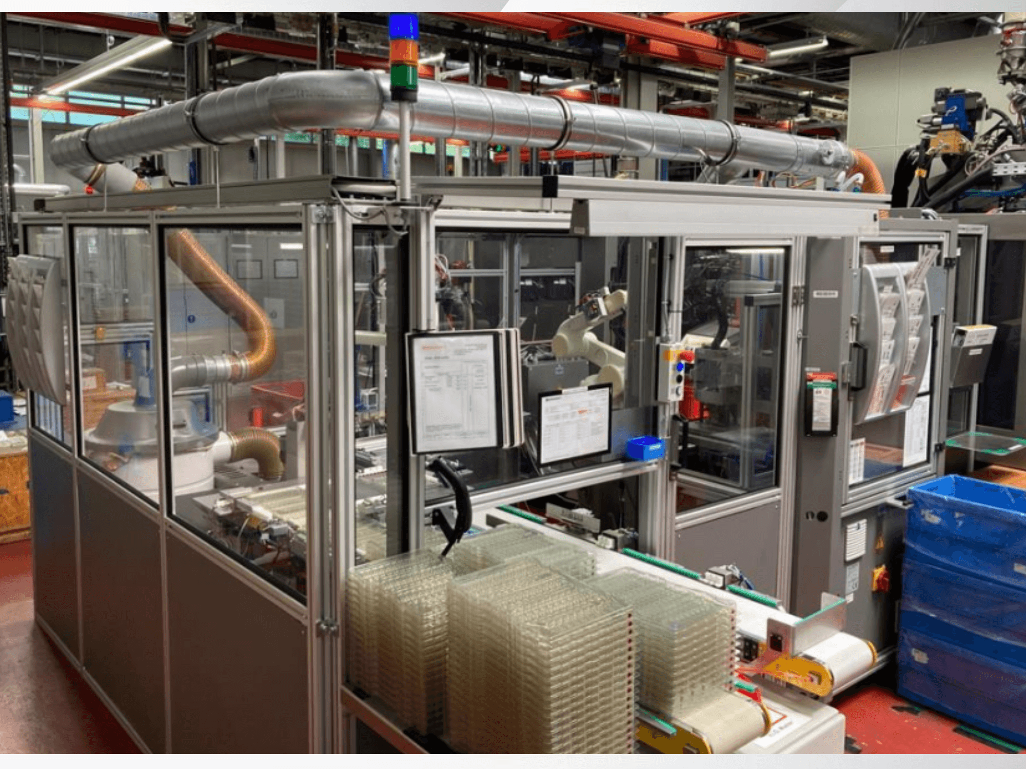 Frontvy av en automatiserad maskin med metallrör, kontrollpanel och staplade behållare av klar plast på ett fabriksgolv.