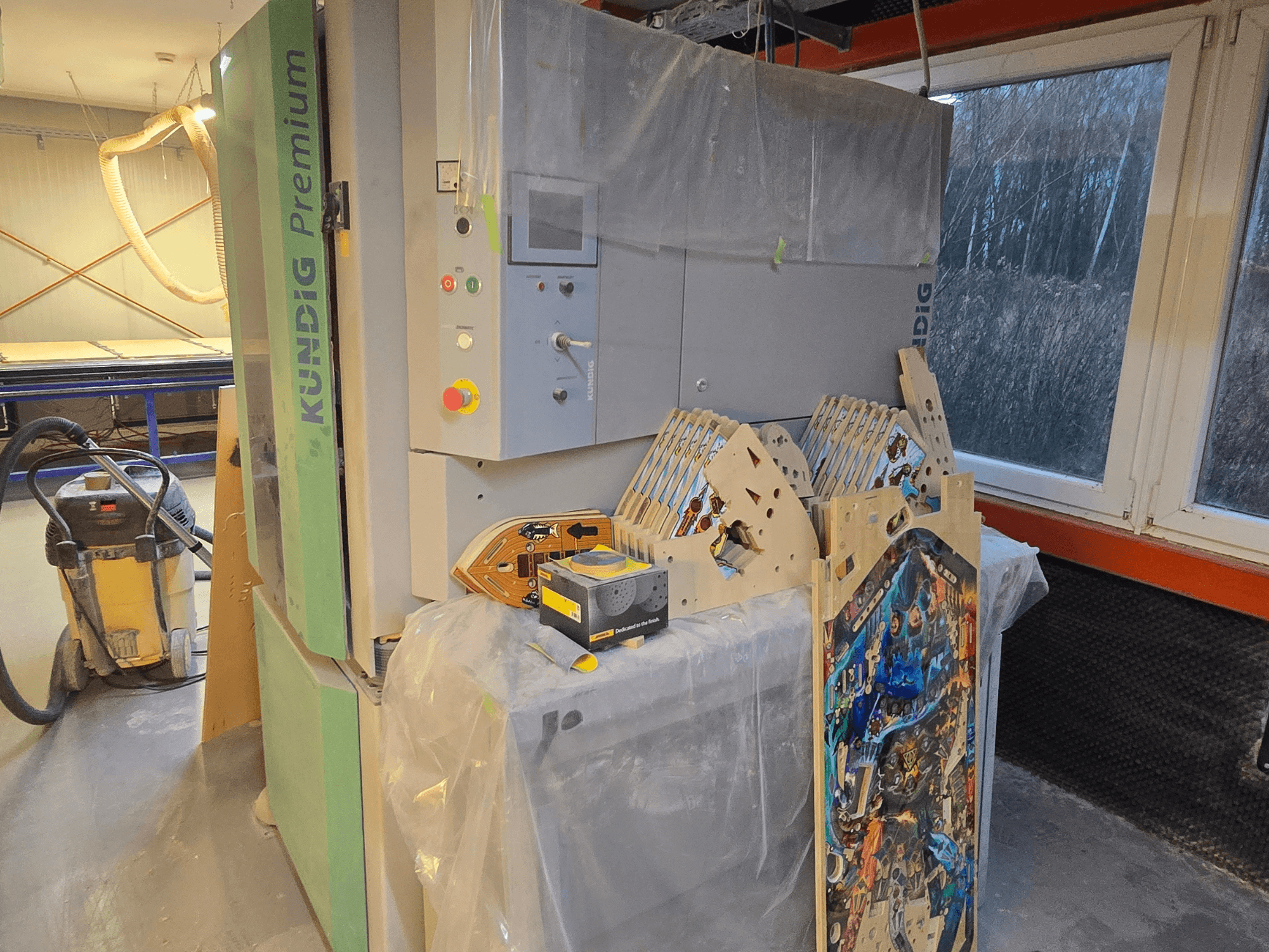 Kundig Premium-1 1100-C bandslip i verkstad, med kontrollpanel och trästycken, sidovy, med dammsugare i bakgrunden.