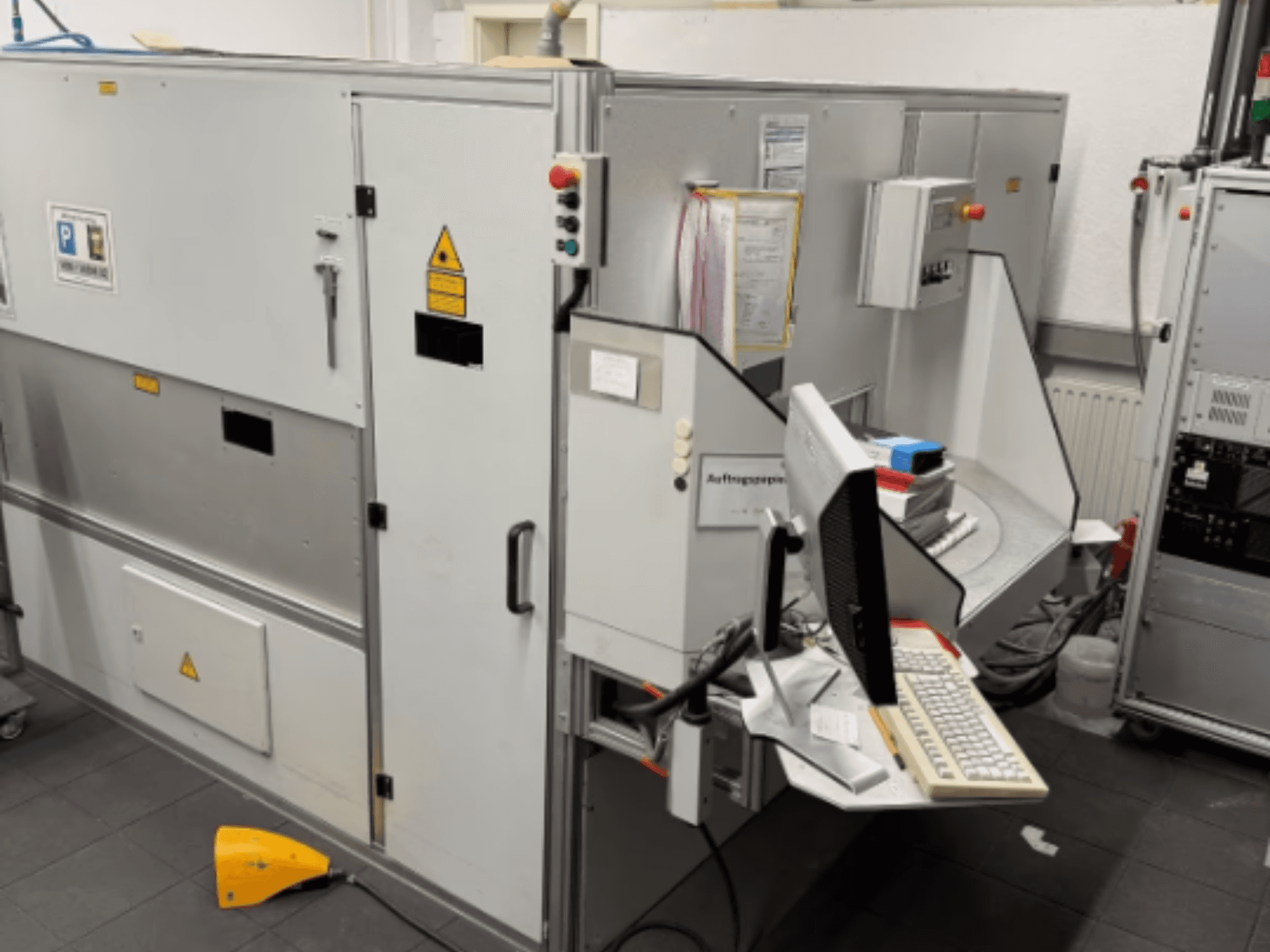 Industrimaskin med datorterminal, vy från sidan; inkluderar kontrollknappar och varningsskyltar.