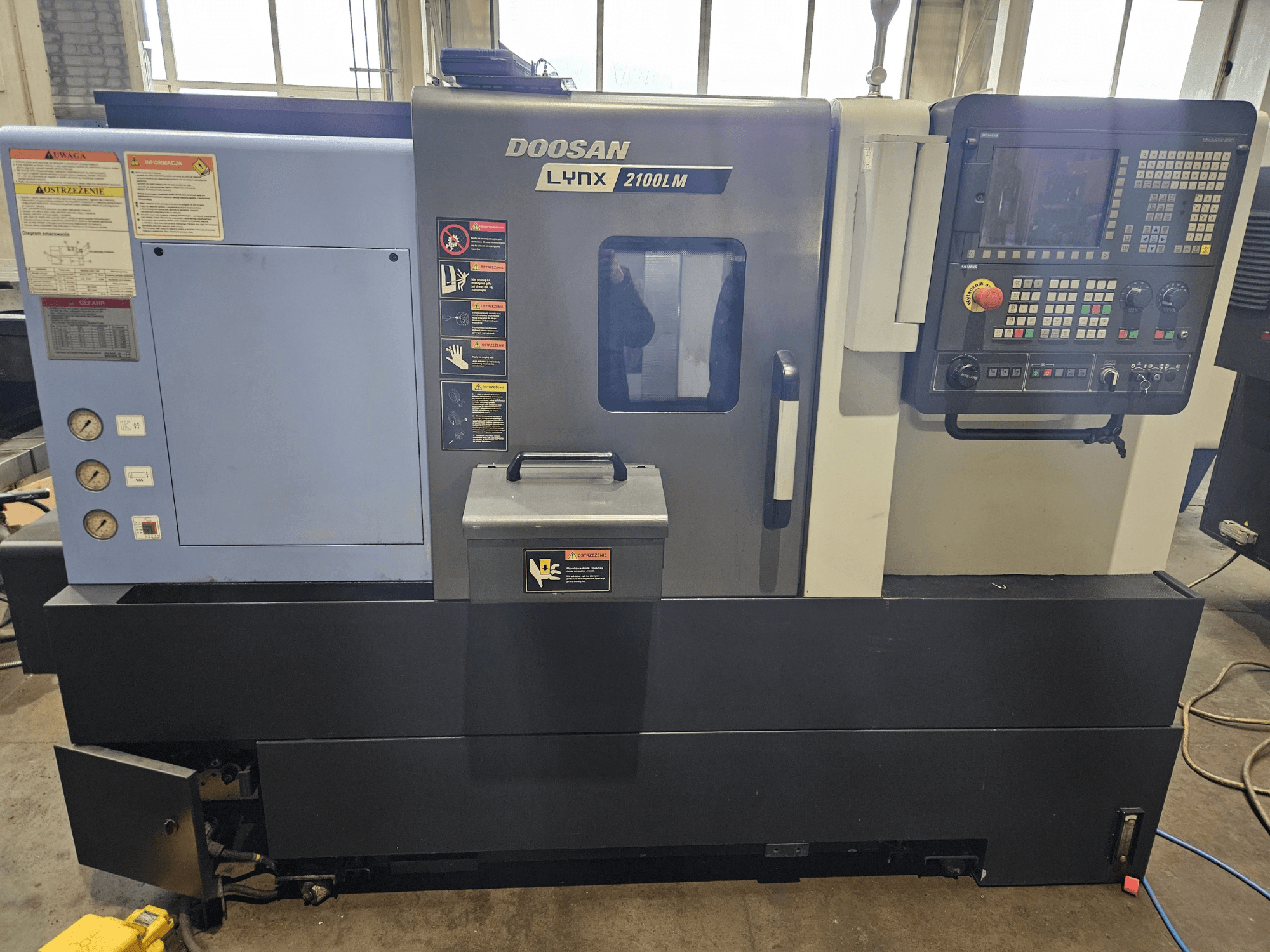 Doosan Lynx 2100LM CNC-svarv, framifrån, med kontrollpanel, lufttrycksmätare och säkerhetsetiketter synliga.