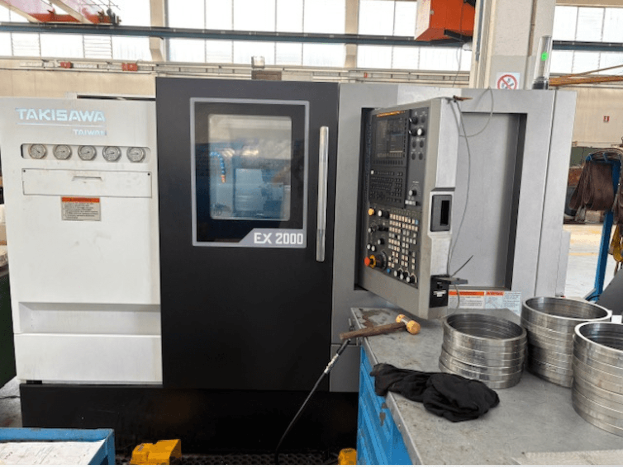 Takisawa EX 2000 CNC-maskin, framifrån, med kontrollpanel, mätinstrument och metalldelar på ett verkstadsbord.