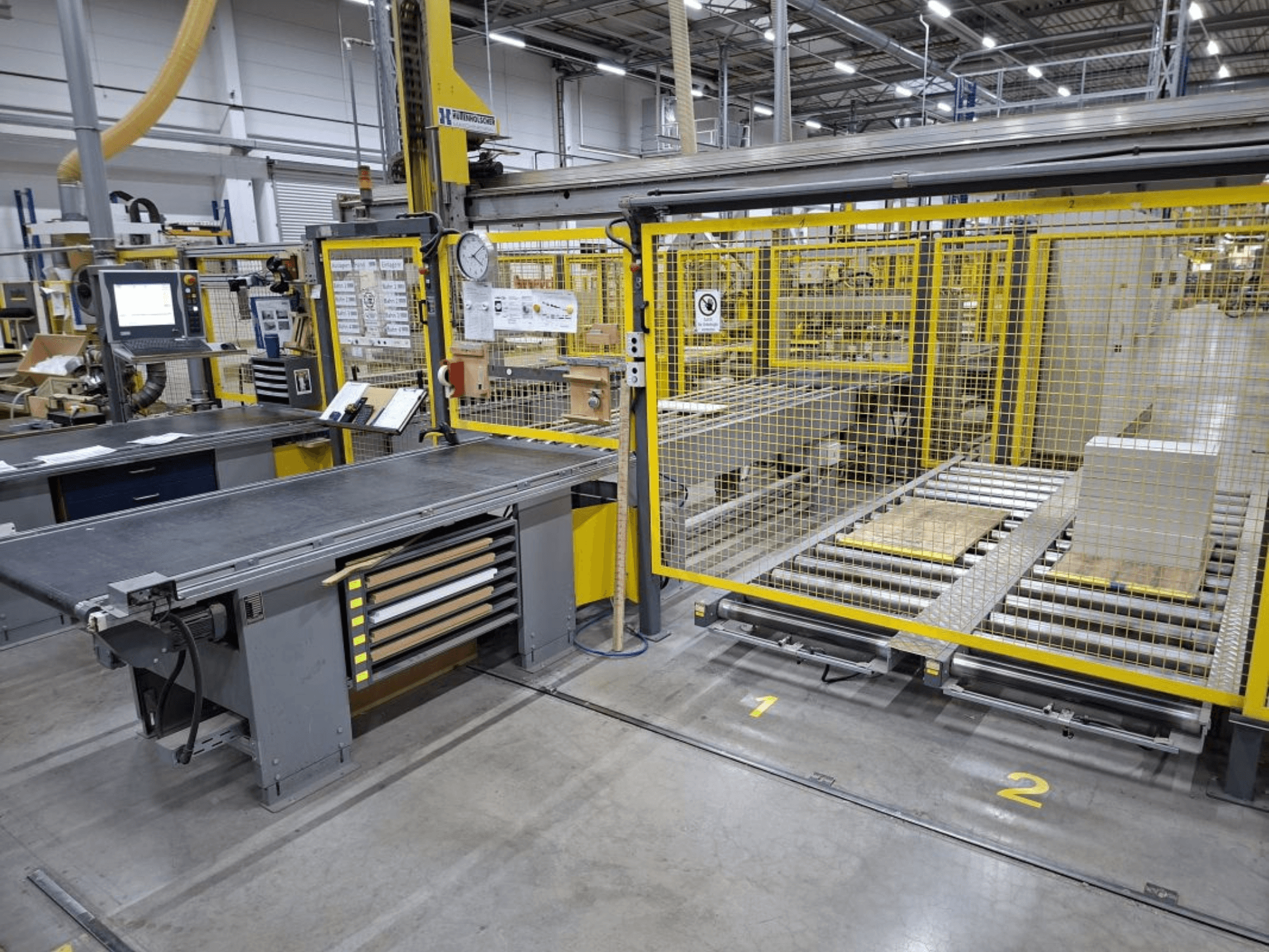 Industriellt arbetsutrymme med transportörsystem och kontrollpanel; sidovy med maskiner och säkerhetsstängsel.