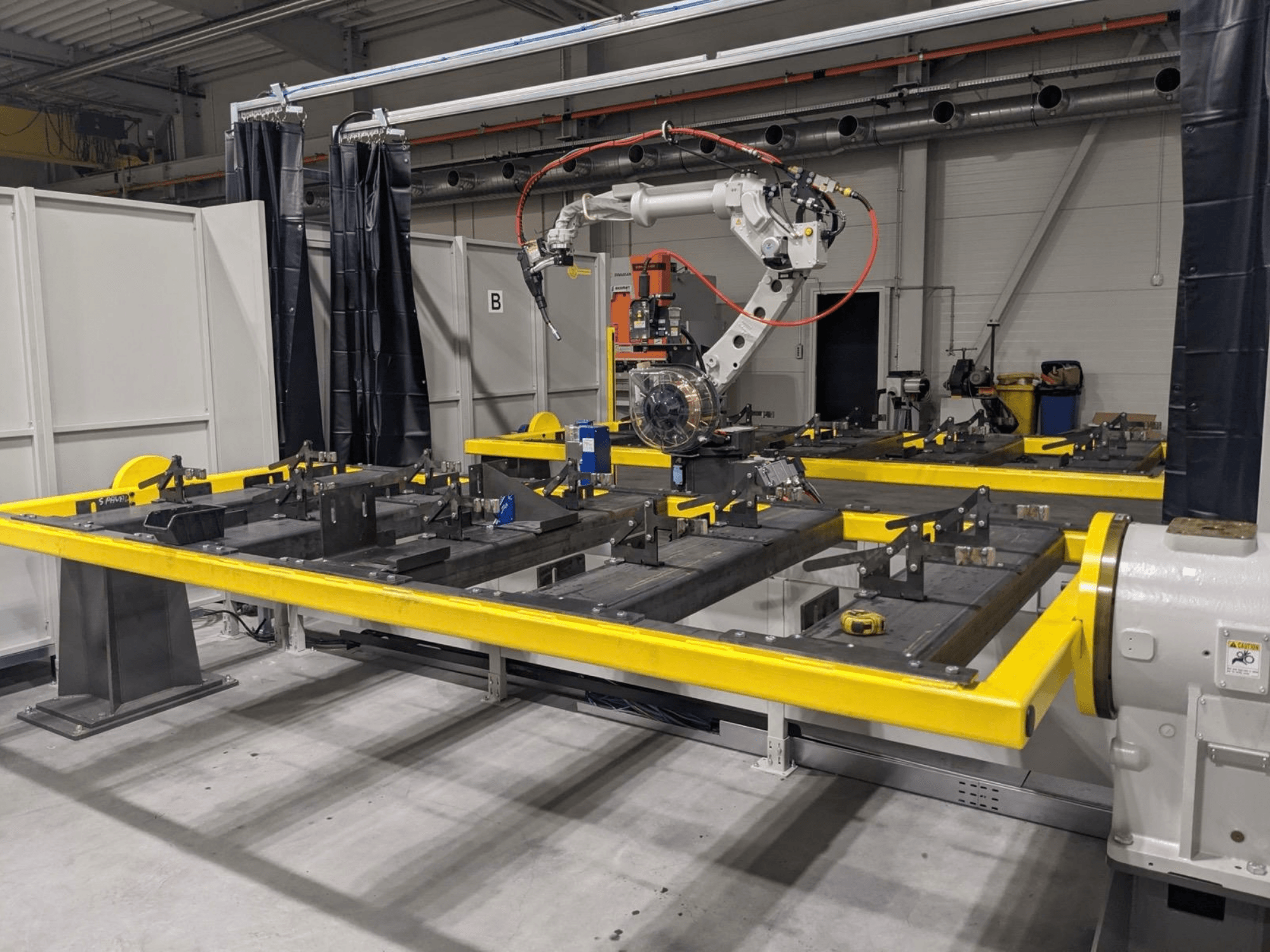 Framifrån sett: PANASONIC TAWERS TM-2000WG3-robotarm placerad ovanför ett gult arbetsbord med metalldelar som ska monteras.