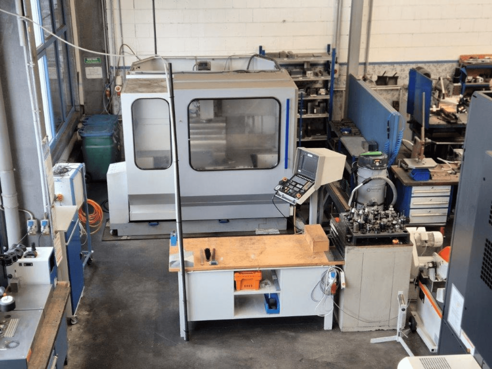 CNC-fräsmaskin i en verkstad, framifrån, med kontrollpanel, arbetsyta och verktyg synliga i bakgrunden.