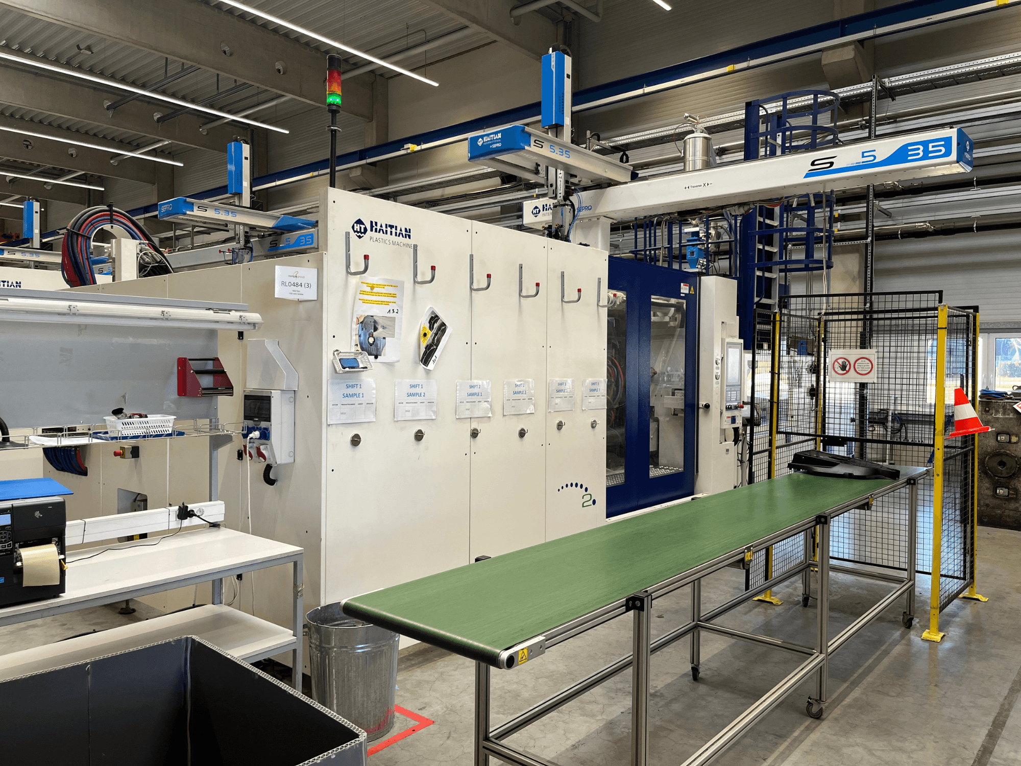 Haitisk S5-35 plastmaskin i en verkstad; grönt transportbord, säkerhetsbarriärer och kontrollpanel synliga framifrån.