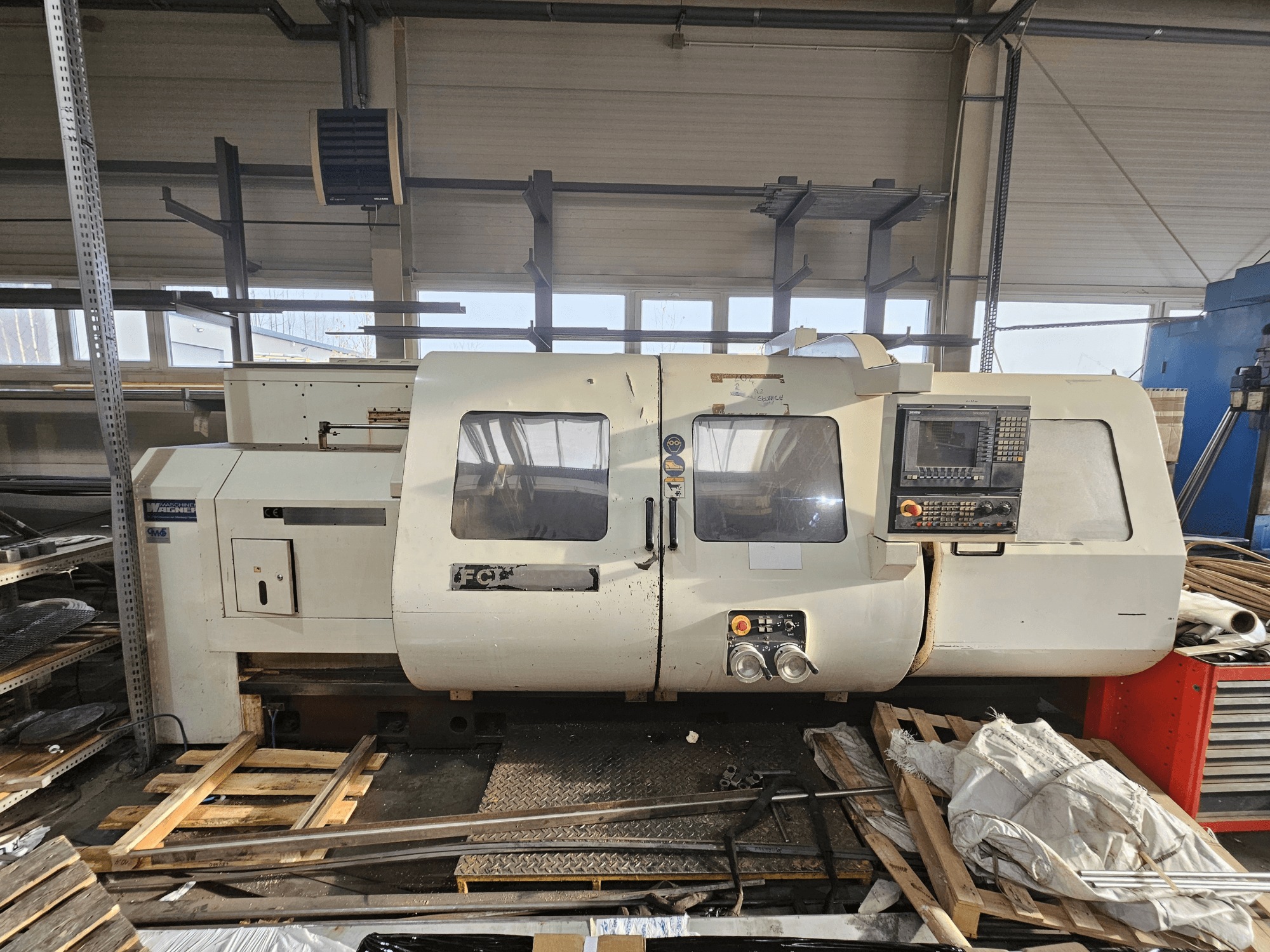 Wabeco CNC-svarv, sidovy, med kontrollpanel, fönster och arbetsområde; maskiner på träpallar i verkstadsmiljö.