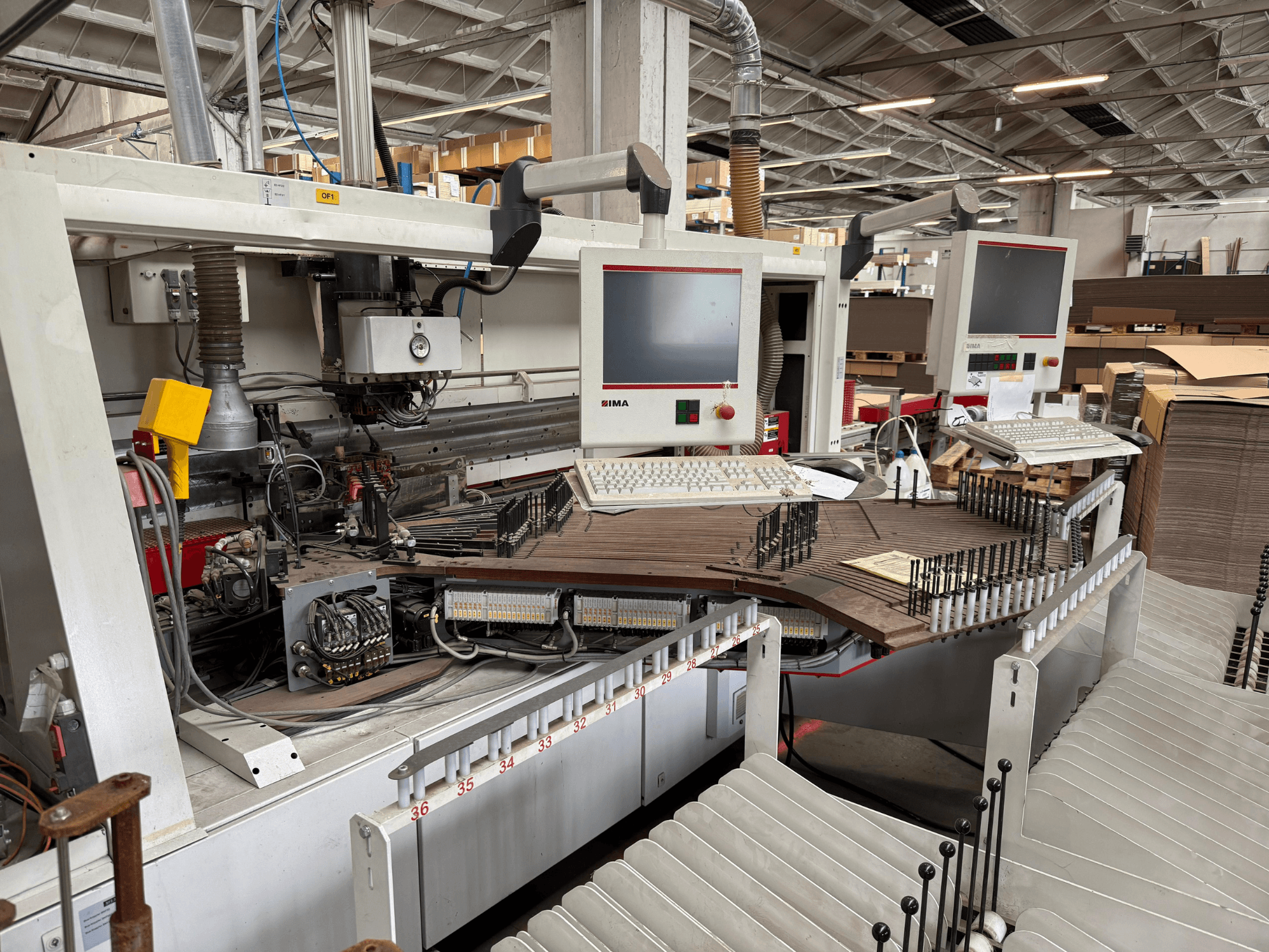 IMA-maskin för precisionsbearbetning av trä med dubbla bildskärmar och kontrollpaneler, framifrån med transportör och verktygsområde.