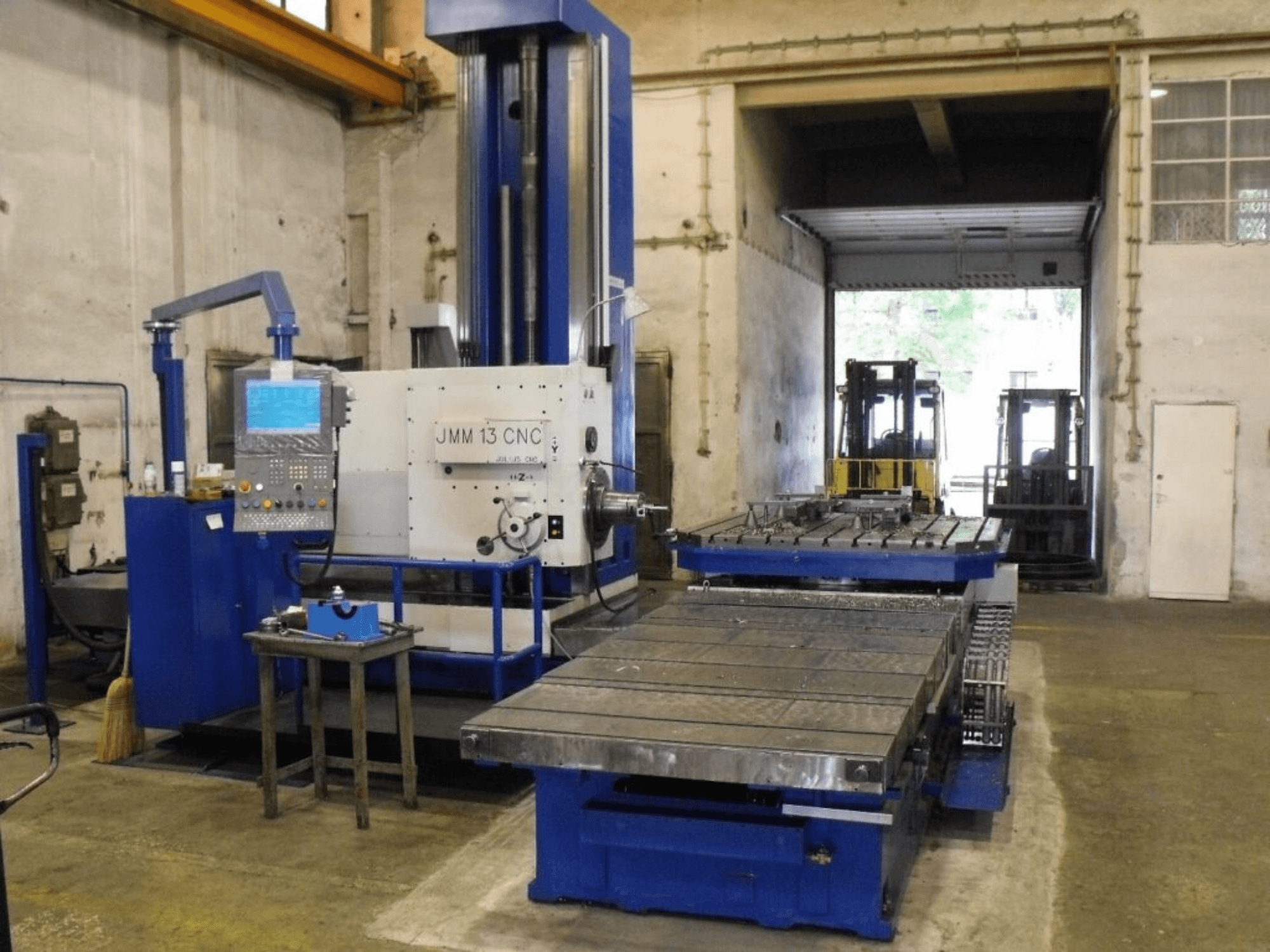 JMM 13 CNC-fräsmaskin i en verkstad, framifrån, med kontrollpanel, stort arbetsbord och industriell bakgrund.