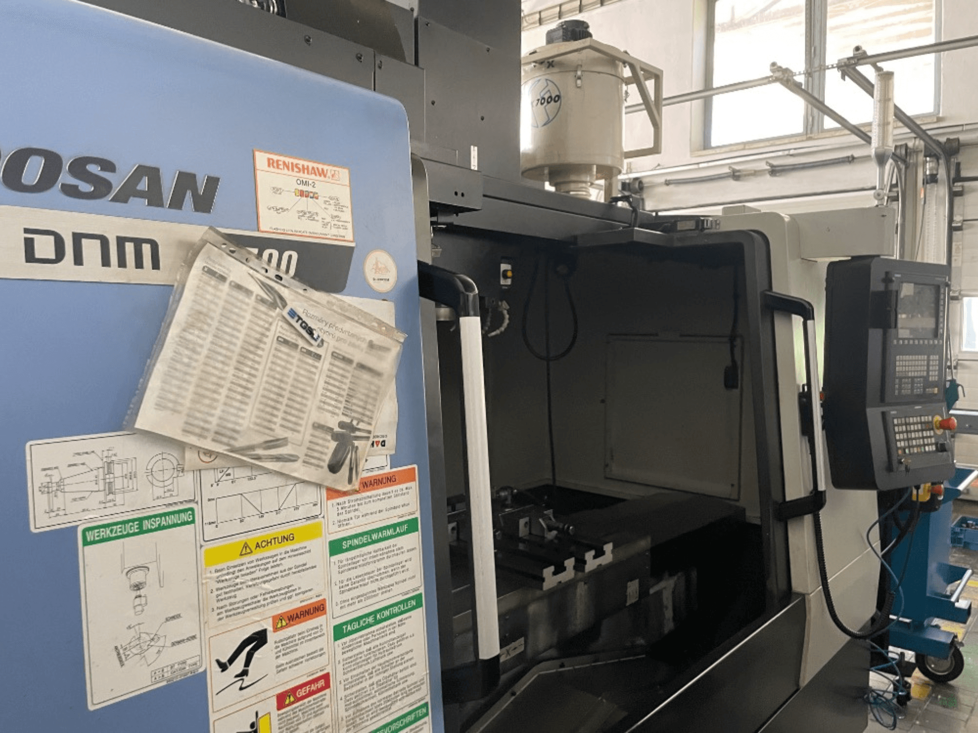 DOOSAN DNM 5700 vertikalt bearbetningscenter, framifrån, visar kontrollpanel, säkerhetsetiketter och maskindetaljer.
